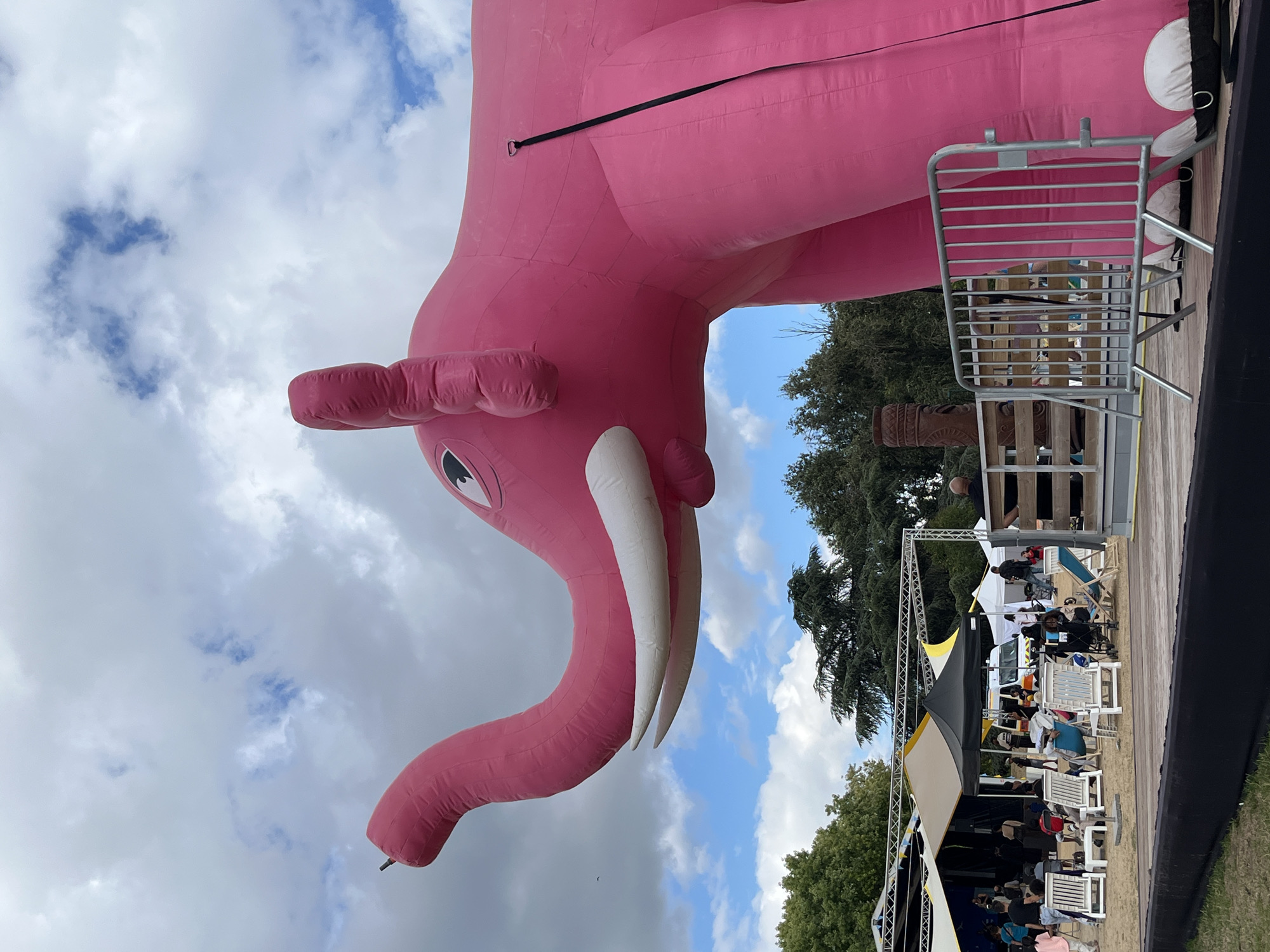 An extremely large, bright pink inflatable elephant is prominently featured in the foreground of the image, dominating the right side. Its trunk is raised, and its tusks are visible. The elephant appears to be part of an outdoor event, possibly a festival or public gathering. In the background, several tents, stages, and a white van are visible. People are scattered throughout the scene, some sitting in white lounge chairs. The environment appears to be a park or open field, with trees and a cloudy sky above. The location is identified as Bobigny, France.