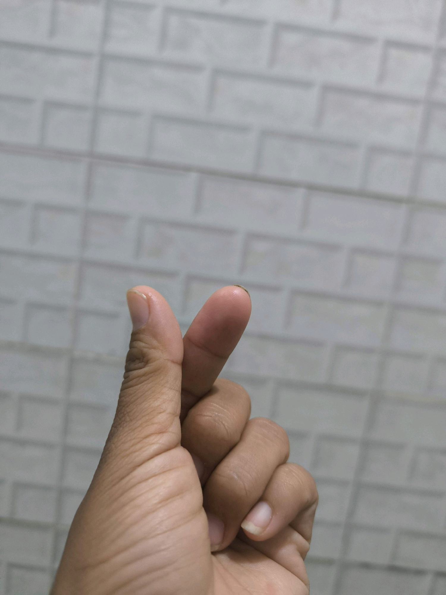 A close-up shot shows a person's hand forming a "finger heart" gesture against a textured, tiled background. The hand, with its skin tone, is positioned in the lower half of the frame, with the thumb and index finger touching to create a heart shape. The background is a muted, light-colored tile pattern, possibly found in a bathroom or kitchen. The lighting is soft, casting subtle shadows on the hand. There are no other people or objects visible in the frame. The image appears to have been taken indoors in a domestic setting. No specific location in Tabanan, Indonesia, can be identified from the visual information provided.