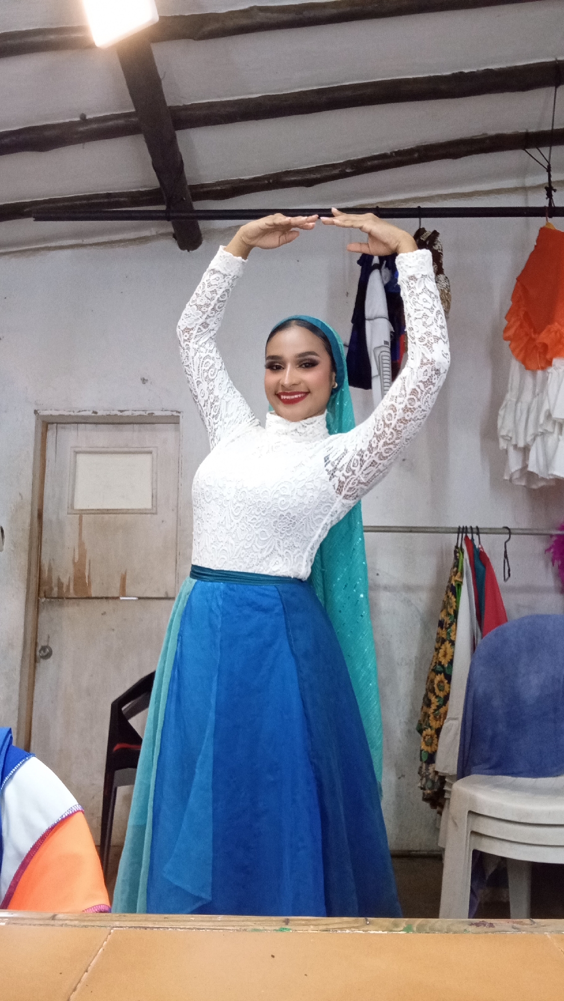 The image depicts a young adult female posing indoors in Porlamar, Venezuela. She stands centrally, facing the viewer directly with a broad smile. Her dark hair is pulled back, covered by a teal/aqua headscarf that drapes over her left shoulder and features subtle shimmer. She wears a long-sleeved, high-necked white lace top and a flowing blue skirt composed of multiple layers in varying shades of blue, secured by a dark teal waistband. Her arms are raised above her head, forearms parallel to the ceiling, with hands held close together, palms facing each other. Her face is adorned with prominent eye makeup and red lipstick.
The background reveals an utilitarian indoor space. To the left, a light-colored wooden door with peeling paint and a rectangular inset panel is visible. Above, the white ceiling is traversed by several dark wooden beams. A bright rectangular light fixture is partially cropped in the upper left corner. To the right, a metal clothing rack holds various garments, including a white ruffled item with orange trim, and a garment displaying a sunflower pattern. A vibrant orange ruffled skirt hangs partially cropped on the far right. A dark chair with a red seat is partially visible behind the woman on the left, and a white plastic chair with a blue fabric draped over it is on the far right. The immediate foreground is occupied by a flat, light brown surface, possibly a table or barrier.