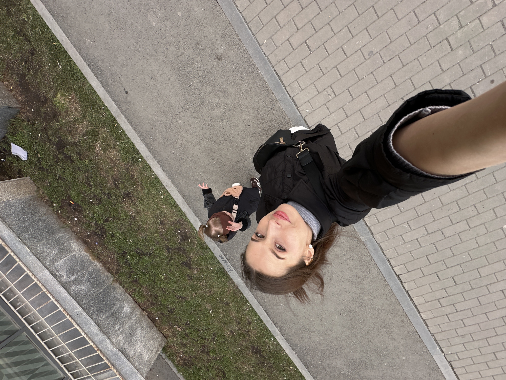 A downward-angled selfie shows a woman and a toddler on an outdoor sidewalk in Yekaterinburg, Russia. The woman, wearing a black jacket over a gray turtleneck, holds the camera. The toddler, wearing a black jacket, brown shoes, and a brown head covering, is to the woman’s left. The sidewalk is divided between pavement and brick. To the left of the pavement is a grassy area littered with debris. A building facade is visible on the far left.