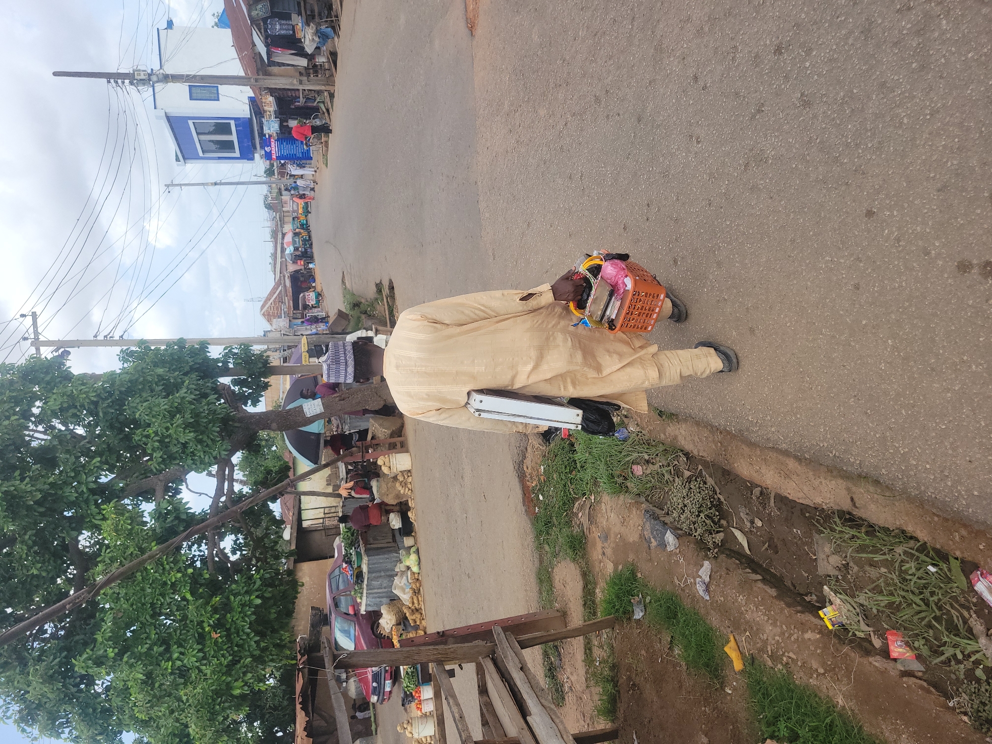 A man in traditional Hausa attire walks along a paved road in Jos, Nigeria. He carries a plastic shopping basket filled with various items, including what appears to be food and personal belongings. A metal briefcase is tucked under his arm. The road is surrounded by a mix of residential and commercial buildings, some with corrugated metal roofs. A wooden structure partially obscures a view of a red car. There is a tree with dense green foliage on the left side of the road. A narrow, overgrown drainage ditch runs parallel to the road. Power lines crisscross the sky in the background, where the sky is overcast. Other pedestrians and market stalls can be seen further down the road.