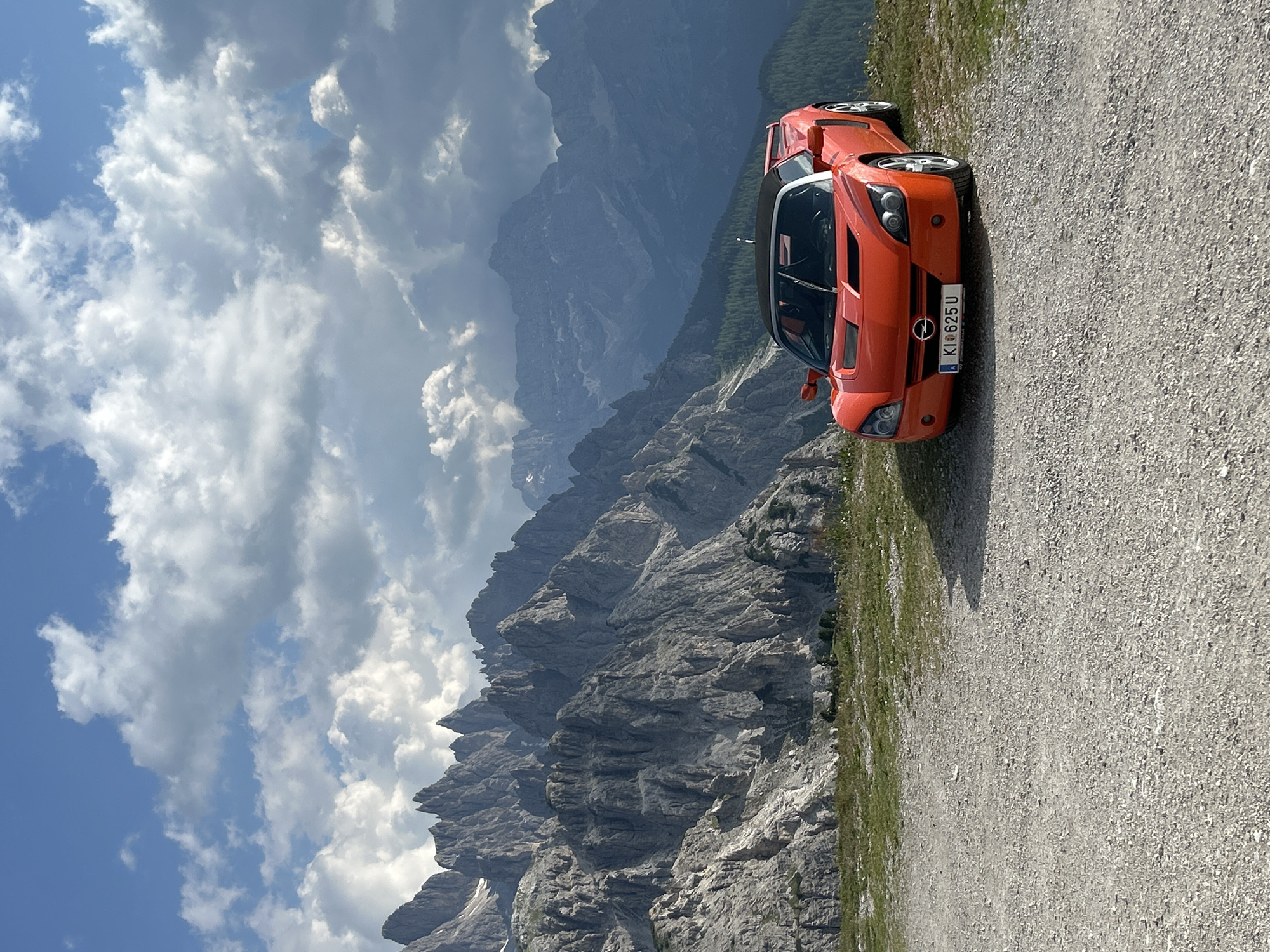 The image shows an orange sports car parked on a gravel road with a mountainous landscape in the background. The car, which appears to be an Opel GT, is facing to the right and slightly away from the camera. Its convertible top is down, revealing the interior. The license plate reads "KI 625 U".
The background consists of rugged, gray mountain peaks under a blue sky dotted with white clouds. The sun appears to be shining, creating a bright atmosphere. The mountains are steep and rocky, with some sparse vegetation visible on the lower slopes. The road the car is parked on is a light-colored gravel surface with a strip of green grass along the edge leading to the mountains.
The scene is a scenic vista, likely in a mountainous region. The time of day seems to be daytime, given the bright lighting. The weather appears to be clear and sunny, with some clouds adding visual interest to the sky. There are no people visible in the image. The overall impression is one of adventure and enjoyment of the outdoors, with the sports car adding a touch of excitement to the dramatic natural setting.