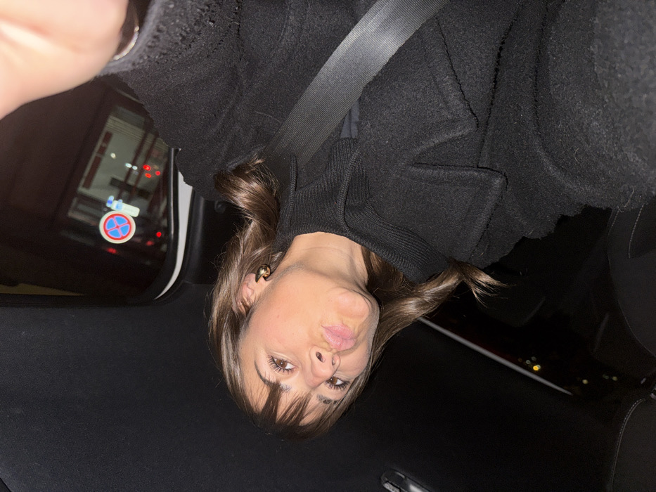 A woman with brown hair and bangs is inside a car, wearing a black coat and a seatbelt. She has gold hoop earrings and is pouting her lips at the camera. A sign with a red and blue circle and a line through it is visible through the car window. The interior of the car is mostly black, and the image appears to be taken at night or in a dimly lit environment.