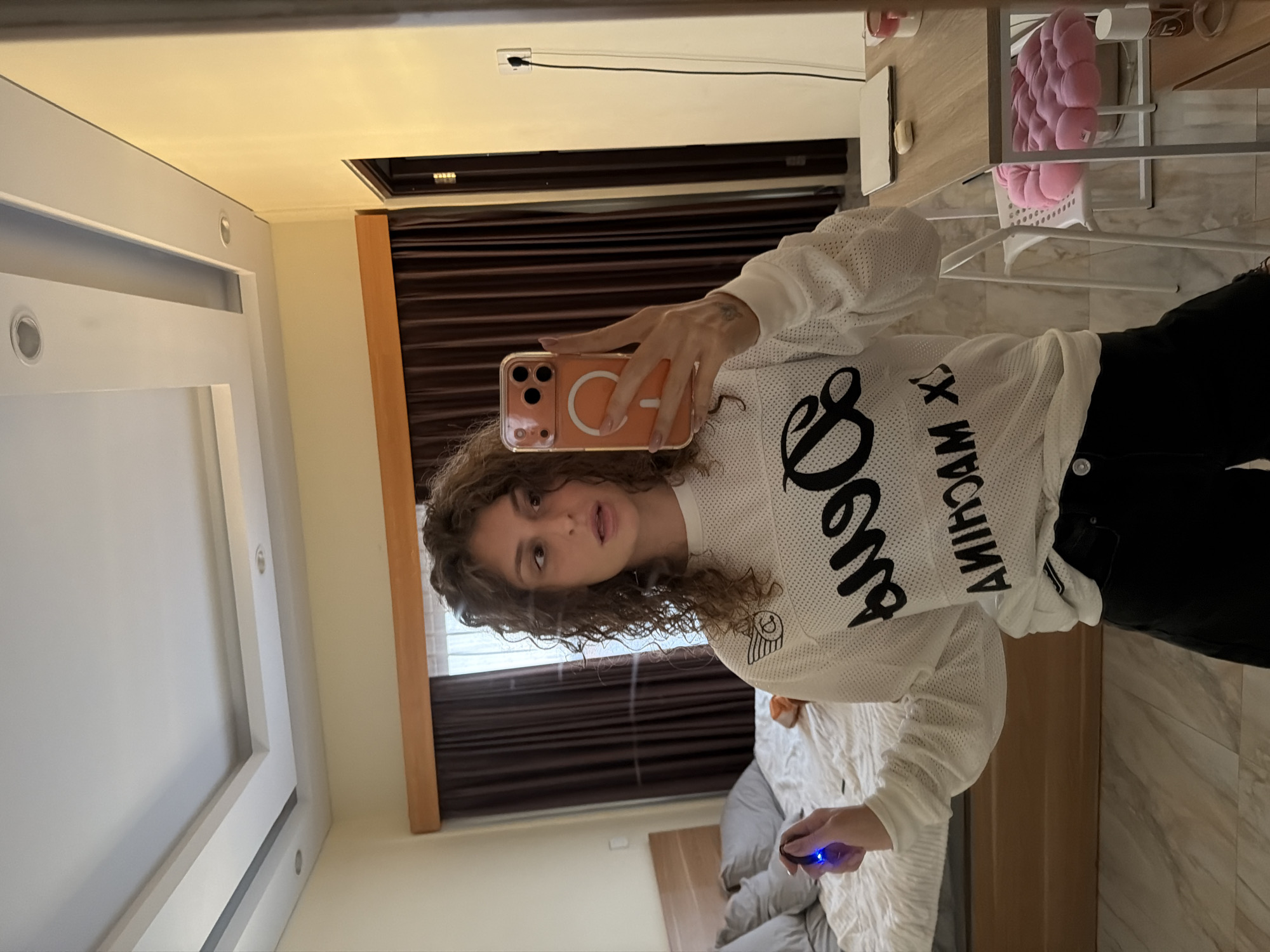 A young Caucasian female is taking a mirror selfie in a bedroom in North Kuta, Indonesia. She has curly brown hair and is wearing a white long-sleeved shirt with black lettering and black pants. She is holding an orange-cased smartphone in her right hand and a small blue object in her left hand. The room features a bed with pillows and a dark curtain in the background, a wooden headboard, and a patterned floor. The lighting suggests it is daytime.