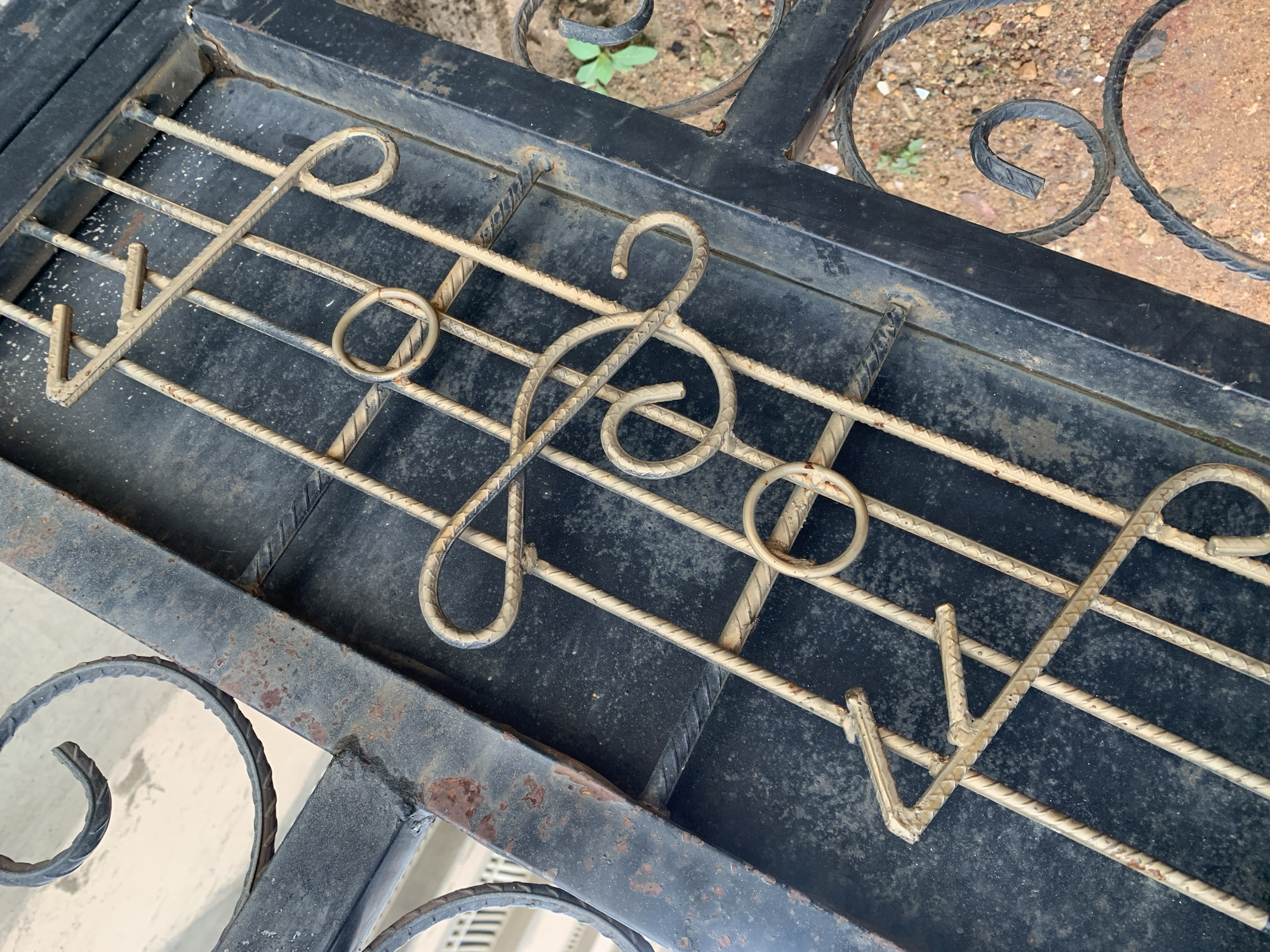 This image shows a close-up of a black metal gate with gold-colored wrought iron decorative elements. The decorative elements are fashioned to resemble musical notation, specifically a treble clef and what appears to be musical notes. The gate is weathered and shows signs of rust in some areas, particularly along the edges of the black metal frame. The background is out of focus, but appears to be a sandy or dirt ground with some small green plants visible. The scene suggests an outdoor environment. The location is identified as Kabong, Nigeria.