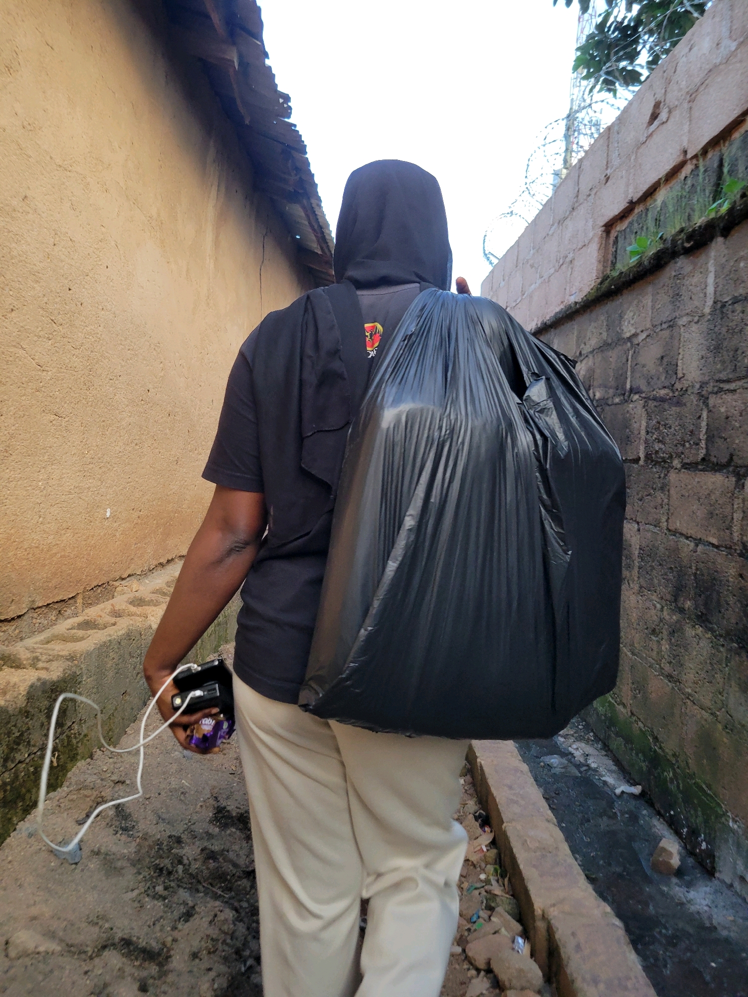 A person wearing a black hoodie and light-colored pants walks away from the camera, carrying a large black trash bag on their back. They are also holding a black electronic device with a white cord and a small wrapped item in their right hand. The person is walking down a narrow dirt path between two walls. The wall on the left is made of tan-colored stucco, and the wall on the right is constructed of gray cinder blocks. A drain runs along the base of the right wall, containing debris and murky water. Barbed wire is visible along the top of the right wall, suggesting a secure or fenced area. Overhanging trees and foliage are visible in the upper right corner. The scene appears to be in an outdoor urban or semi-urban environment within the city of Jos, Nigeria, possibly a less developed residential area or alleyway. The lighting suggests daytime.