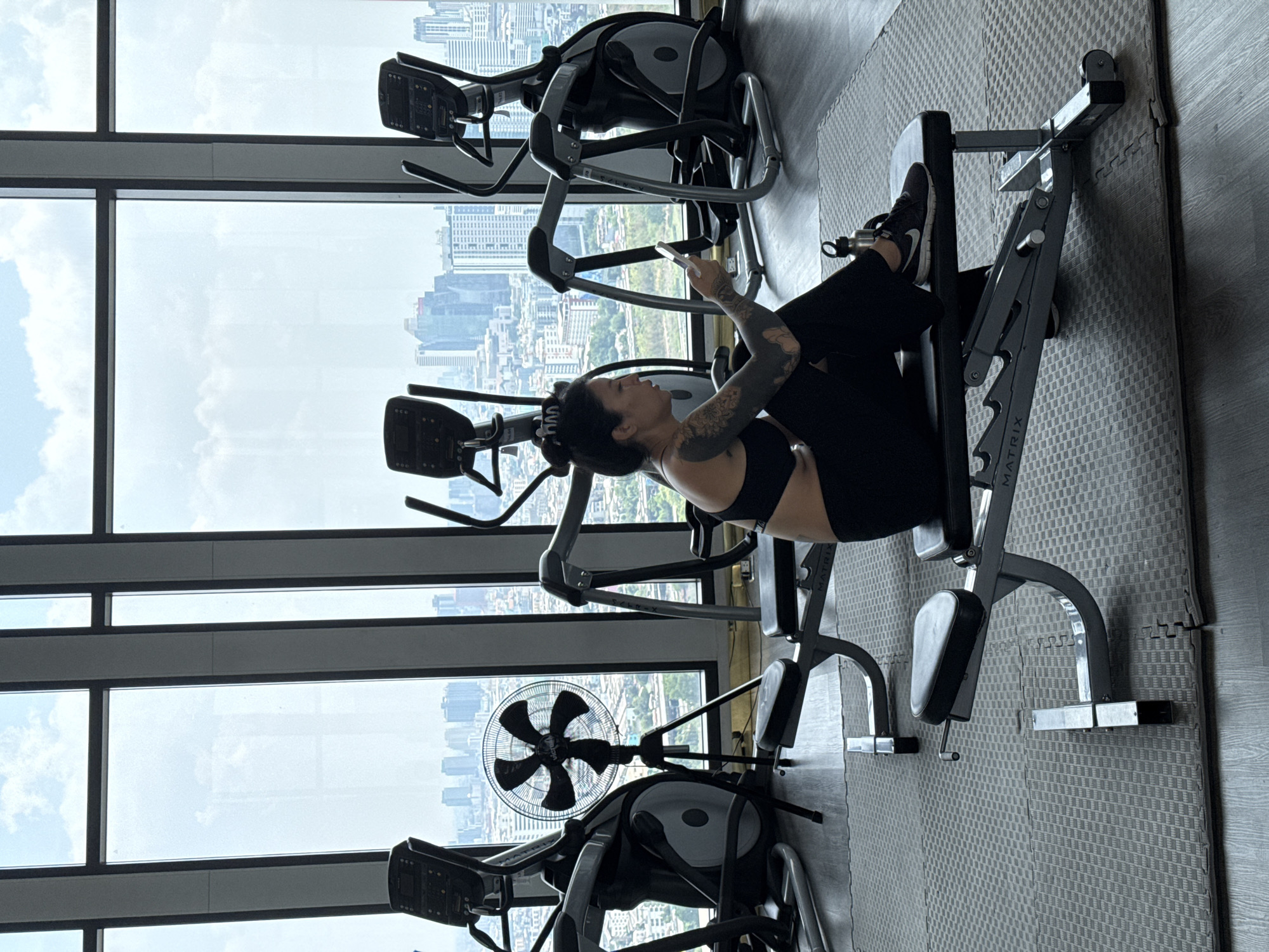 A sideways, medium shot shows a woman using a leg-press machine in a gym. The woman, who is facing left, is wearing a black sports bra and black leggings. She has a high ponytail and has tattoos on both arms. She is looking down at a cell phone in her hands. The leg-press machine is black metal with black padded seats. A treadmill is to the woman's left. Floor to ceiling windows surround the gym equipment. A cityscape of Bangkok, Thailand is seen through the windows.