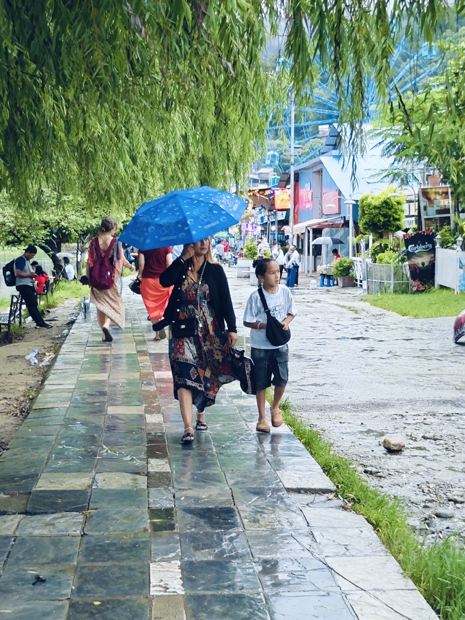 Two people, a woman and a child, walk along a wet, paved path under a large blue umbrella. The woman, in the foreground, wears a patterned dress and a dark cardigan. The child, to her right, wears a light-colored t-shirt and shorts, with a dark bag slung across his chest. Behind them, other pedestrians are visible, some with umbrellas, and a woman in a light-colored dress with a backpack walks away from the camera. The path is bordered by grass on the right and a bank with scattered debris on the left. In the background, buildings with colorful facades line the street, and a Ferris wheel is visible in the distance. The scene appears to be in Pokhara, Nepal, indicated by the distinctive architecture and the presence of a Ferris wheel, a common attraction in tourist areas of the city, especially Lakeside. The overall atmosphere is of a rainy day in a bustling urban environment.