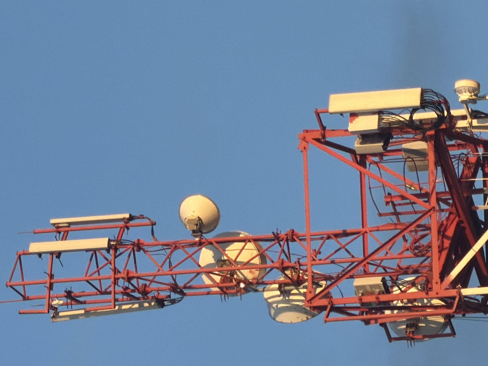 The image shows a close-up view of a red lattice tower, likely a telecommunications tower, against a clear blue sky. The tower is equipped with several antennas and dishes. Multiple rectangular, light-colored antennas are mounted vertically on the tower. There are also several large, circular, white satellite dishes. Some smaller gray equipment boxes are attached to the tower structure, and cables are visible running between them and the antennas. The tower's red metal framework is intricate and shows a crisscrossing pattern of beams. The lighting suggests it is daytime, possibly late afternoon due to the warm tones on some of the equipment. The location is described as Unknown, Unknown, indicating no specific geographical context is available.