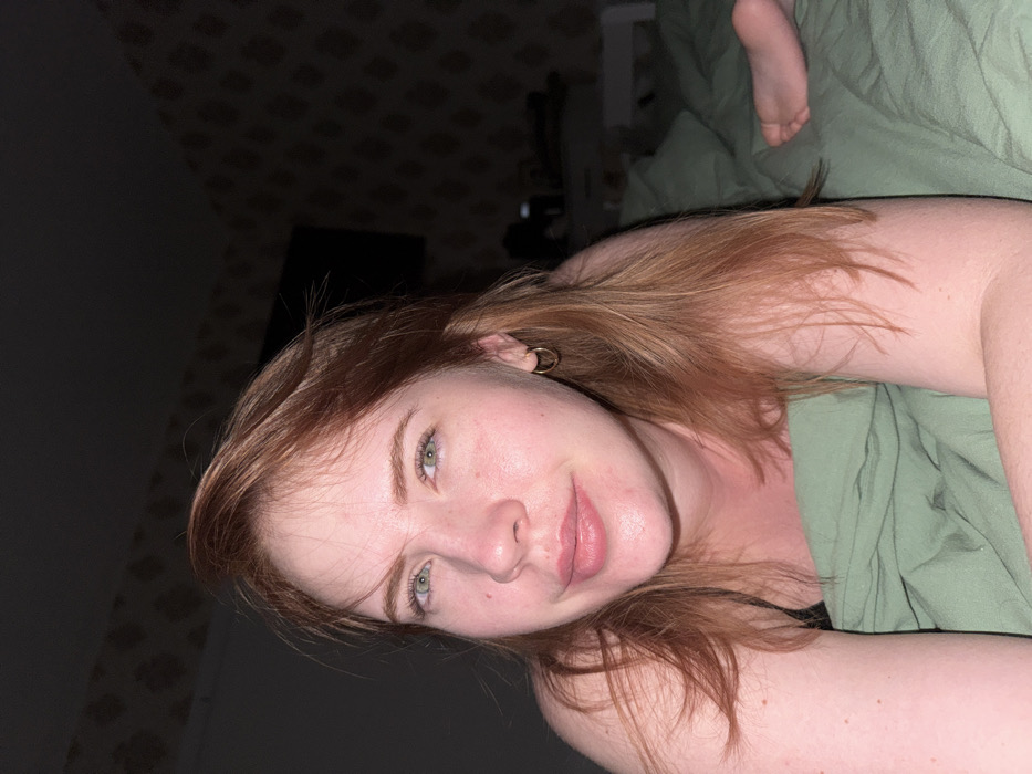 A young woman with reddish-blonde hair and green eyes reclines on a bed with green sheets. Her arms are positioned at her sides. She has fair skin with some visible freckles. She is wearing a small gold hoop earring. The room is dimly lit, with a patterned wall and a television screen visible in the background. A foot is partially visible at the top of the image.