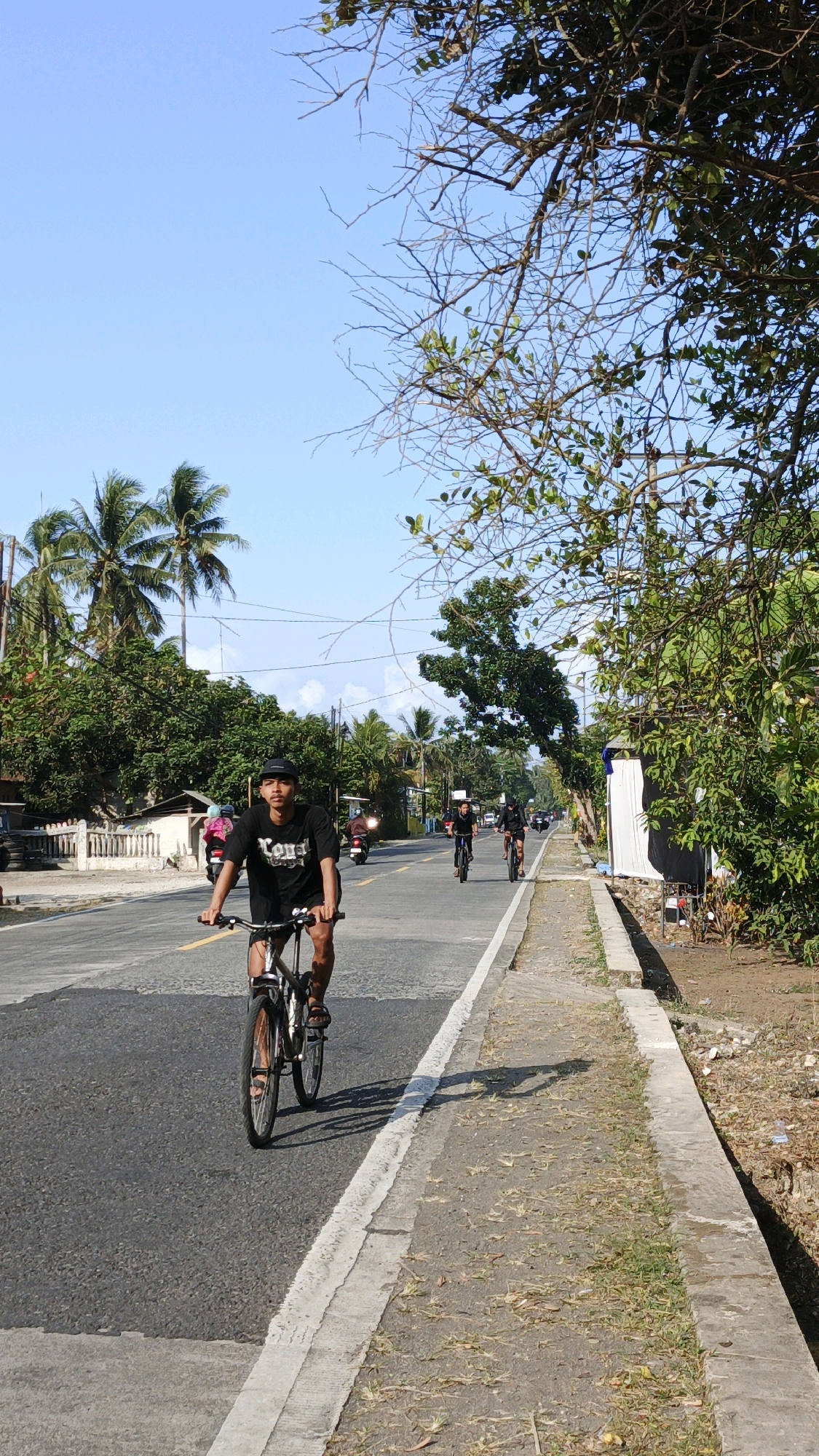 In Cibenda, Indonesia, a sunny day sees a young man, wearing a black t-shirt and shorts, riding a bicycle along the asphalt road. He is positioned on the left side of the road, facing forward with a black cap on his head. Behind him, two other cyclists, one in a black shirt and shorts, the other in a dark top and shorts, are also riding bicycles further down the road. Several motorcycles are visible, one of which appears to be moving ahead of the cyclists. Palm trees and lush green foliage line the sides of the road, with a low white-fenced structure on the left and a concrete sidewalk bordering the right side of the road, which has dry grass and debris. The sky is clear blue, indicating fair weather.