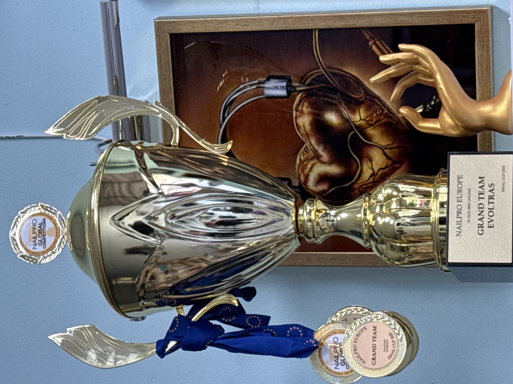 The image depicts a golden trophy, possibly awarded for nail art or beauty competition, with "NAILPRO GLOBAL COMPETITIONS" inscribed on a small medallion attached to it. The trophy also has a blue ribbon and several coins or medallions hanging from it, labeled "NAILPRO EUROPE GRAND TEAM FINAL CUP 2025". In the background, there's a framed picture showing an anatomical heart with tubes and a golden hand pointing towards it. Based on the inscriptions on the trophy, the event might be related to nail art or beauty contests. There's no explicit mention of Burgas, Bulgaria in the image, but the trophy alludes to European competition, indicating a possible location in that region.