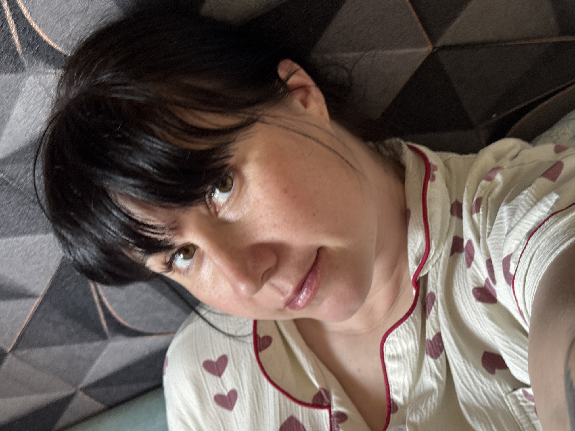 A woman is lying down, wearing a white pajama top with red heart patterns and red piping, her head resting against a gray geometric patterned surface. Her dark hair has bangs. She is looking towards the camera with brown eyes. The location is not evident in the image.