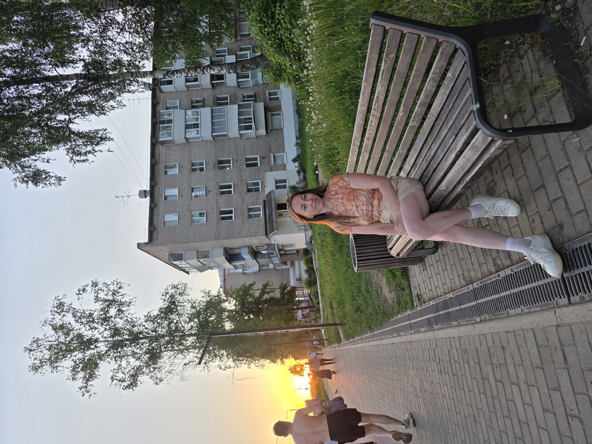 A photograph depicts a woman sitting on a park bench. She is wearing a patterned shirt, shorts, white socks, and white sneakers. Behind her is a multi-story brick apartment building. In the foreground, a paved walkway is visible with two men running away from the setting sun. The setting sun is a vibrant orange. The image was taken in the city of Chaikovsky, Russia.