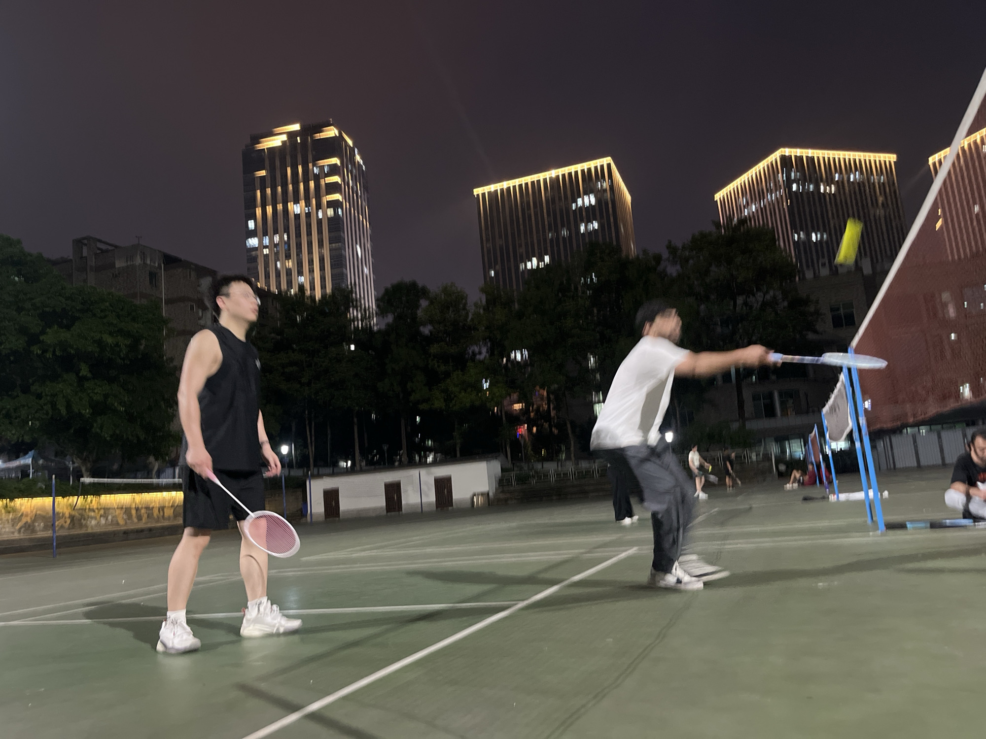 In Yuzhong District, China, at night, two men are engaged in a badminton match on an outdoor court. The court is illuminated, highlighting the green surface with white lines marking its boundaries. A badminton net spans the center of the court.
The man on the left, wearing a black sleeveless shirt and black shorts, stands with a pink and white badminton racket in his right hand. He is wearing white athletic shoes.
The man on the right, dressed in a white t-shirt and dark pants, is in motion, swinging a badminton racket. He is also wearing athletic shoes. A yellow shuttlecock is visible in mid-air between the two players.
In the background, several trees are visible. Behind the trees, two tall, modern buildings with illuminated facades stand out against the dark night sky. The buildings have a grid-like pattern of lights. There are also other indistinct figures and structures in the background, suggesting a public recreational area. The overall atmosphere suggests an evening sports activity.