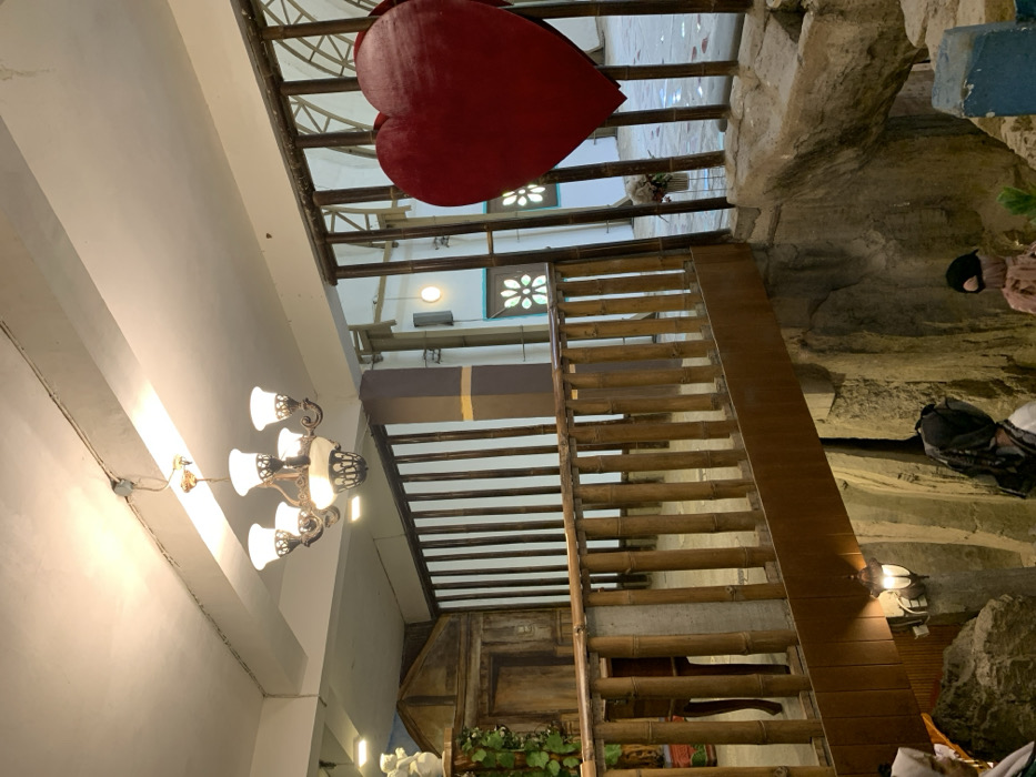 The scene is indoors in a building in Kecamatan Borobudur, Indonesia, located at latitude: -7.606, longitude: 110.18. There is a wooden walkway with bamboo railings. The walkway is suspended over a deep, rocky pit with a blue area in the lower right corner. There is a large red heart-shaped object hanging from the ceiling in the background, surrounded by a latticework structure. A chandelier hangs from the ceiling above the walkway. There is a wooden cabinet in the background and a small table with a wooden top under the walkway. There are several people in the scene, wearing head coverings, but their faces are not visible. The scene is lit by a single overhead light and the chandelier. The environment is relatively dark and appears to be a cave or an underground space.