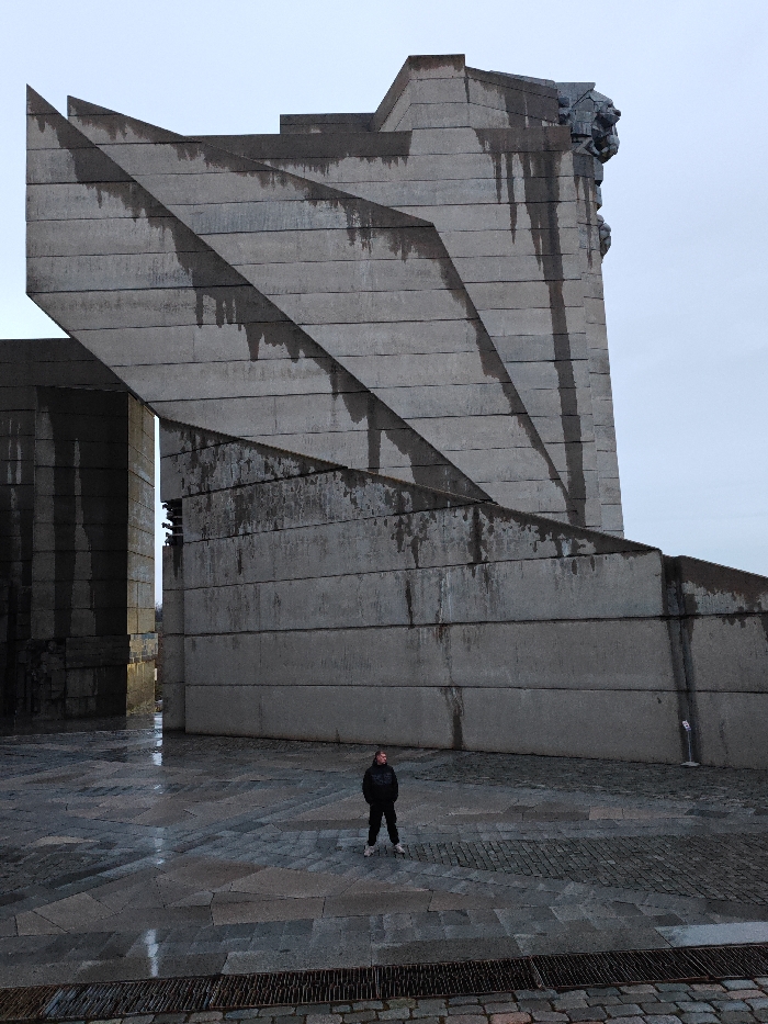 An imposing concrete monument, identified as the Monument to 1300 Years of Bulgaria, also known as the Founders Monument, stands on a plateau overlooking Shumen, Bulgaria. The structure is composed of massive, angular brutalist concrete blocks with visible dark streaks and water stains indicative of weathering. A large, stylized lion's head sculpture is visible on the upper right side of the main concrete mass. In the lower center of the frame, a single individual stands on a wet, patterned stone-paved plaza, facing towards the left side of the towering monument. This individual is dressed in dark clothing, consisting of a black hooded top, dark trousers, and white-soled athletic shoes. The sky is overcast and light grey, contributing to a muted atmosphere. A metal drainage grate runs across the foreground.