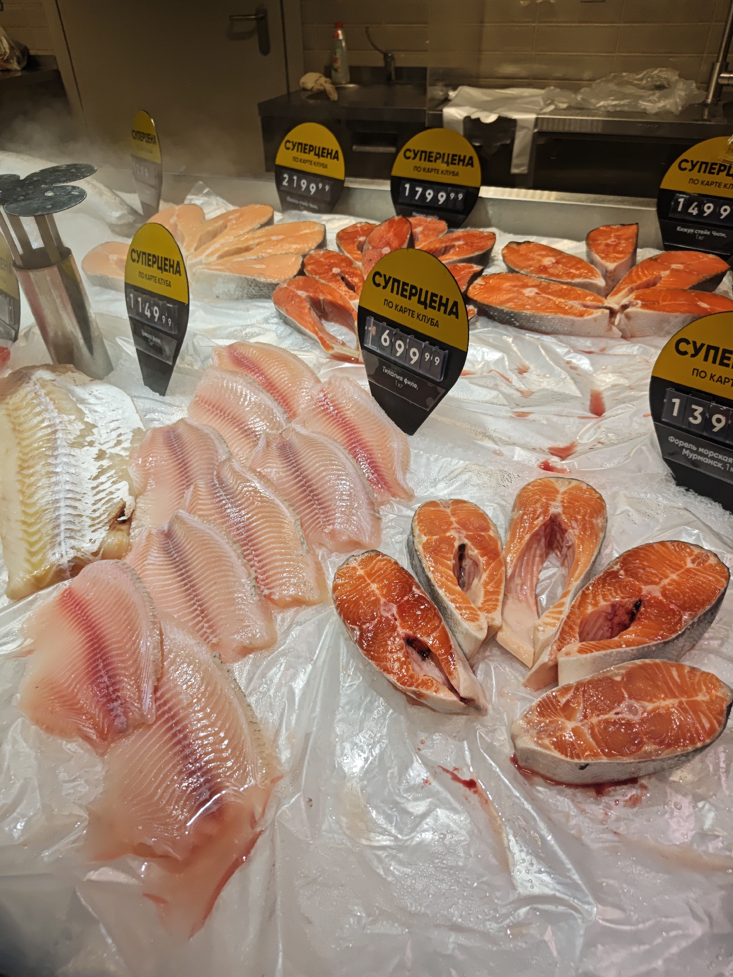 The image shows a fish counter in a store, likely a supermarket or specialty food shop. The fish is displayed on ice, covered by a clear plastic sheet. The fish varieties visible include what appear to be salmon steaks, salmon fillets, tilapia fillets, and another white fish fillet. Price tags, labeled "SUPER PRICE" in Russian ("СУПЕРЦЕНА") and "BY CLUB CARD" ("ПО КАРТЕ КЛУБА"), are stuck into the display next to each type of fish. Price tags indicate items are on sale. Based on the price tags, the location is likely in Russia, possibly Yekaterinburg, as the writing on the price tags is in Russian.