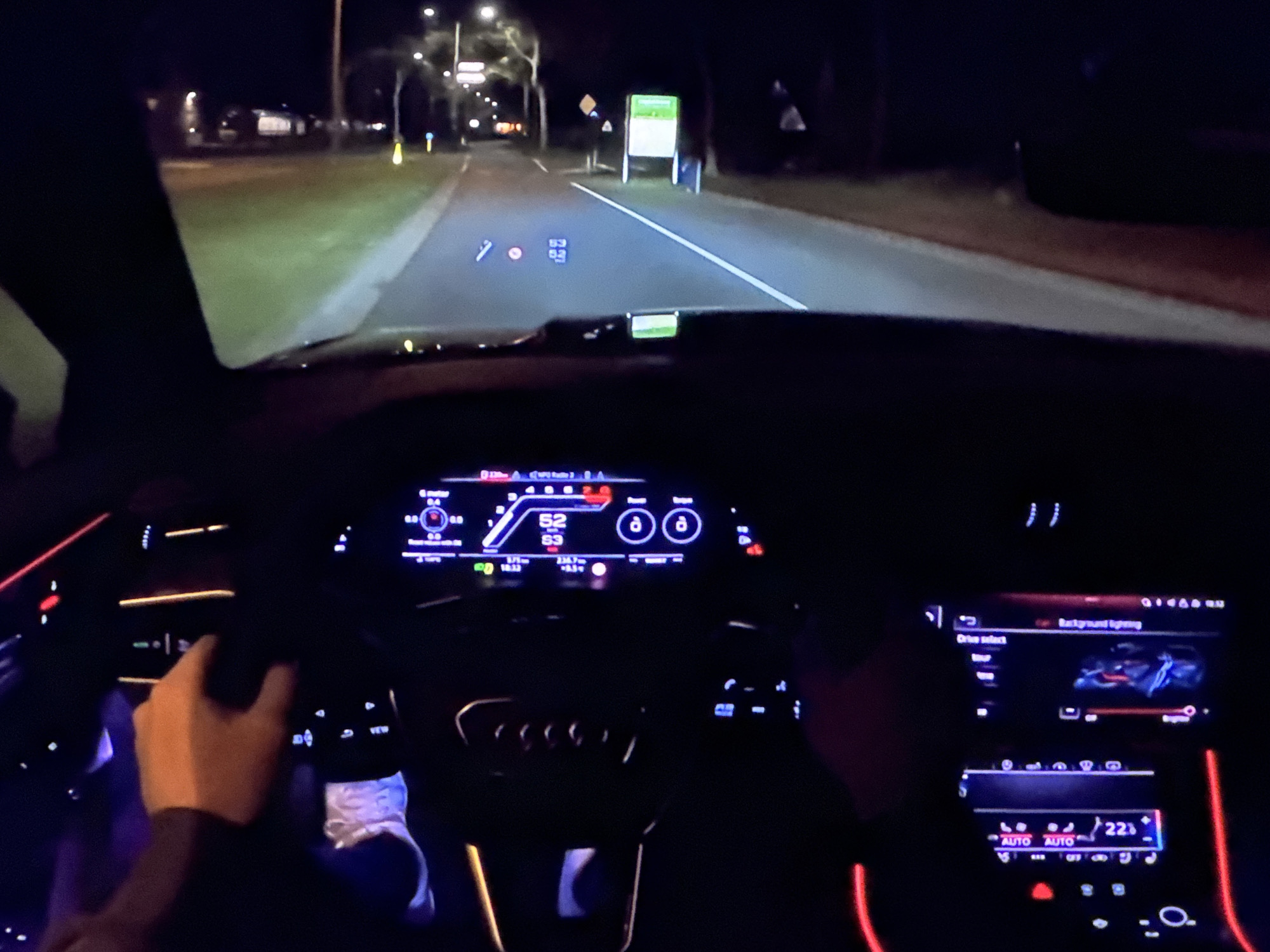 The image shows the interior of a car driving at night on a road that appears to be in or near Bezden, Bulgaria. The driver's hands are visible on the steering wheel, and the car's dashboard displays are illuminated, showing speed, gear selection, and other information. The road ahead is lit by the car's headlights and streetlights, with trees and buildings visible on the sides. The scene depicts a typical night-time drive, with the focus on the car's interior technology and the road ahead.