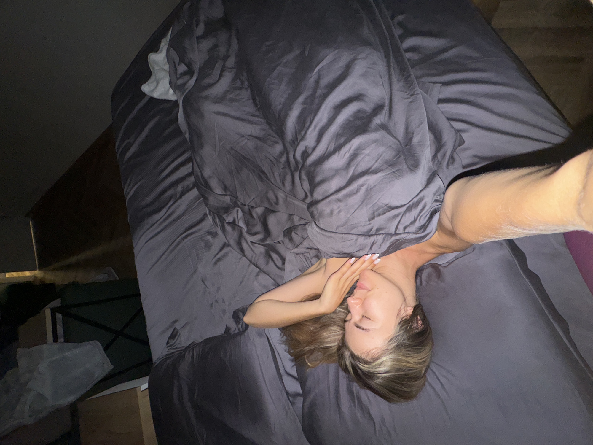 A woman with blonde hair, wearing dark grey sheets, lies on her right side in bed, facing the camera. She is looking up with her eyes closed and her right hand resting on her cheek. Her left arm is bent and her hand is resting on the sheets. The sheets are wrinkled and appear to be made of a soft material. There is a pillow under her head. The room is dimly lit, and the background is out of focus. The image appears to have been taken from a high angle.