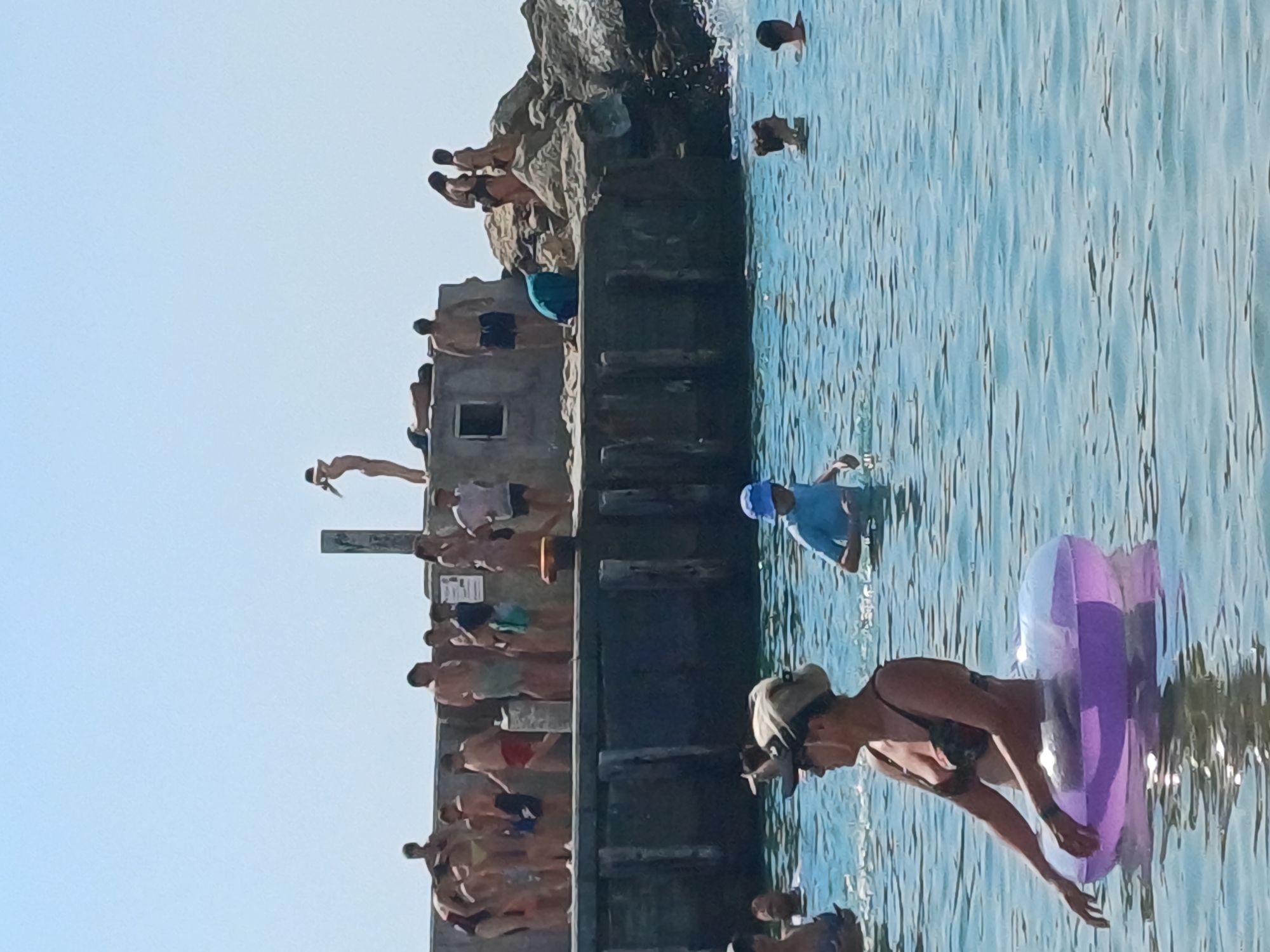 In the city of Varna, Bulgaria, a sunny day at the beach is depicted with clear skies and calm, blue water. In the foreground, a woman wearing a bikini and a hat is partially submerged in the water, holding onto an inflatable ring. Behind her, several other people are in the water, some with their heads visible. In the mid-ground, a group of individuals are gathered on a concrete structure extending into the sea, which appears to be a pier or a breakwater. Some are standing, while others are sitting or lying down. One person is standing at the edge of the structure, seemingly about to jump or dive into the water. The background shows more rocks and the open sea. The overall scene captures a recreational atmosphere typical of a summer day at the seaside.