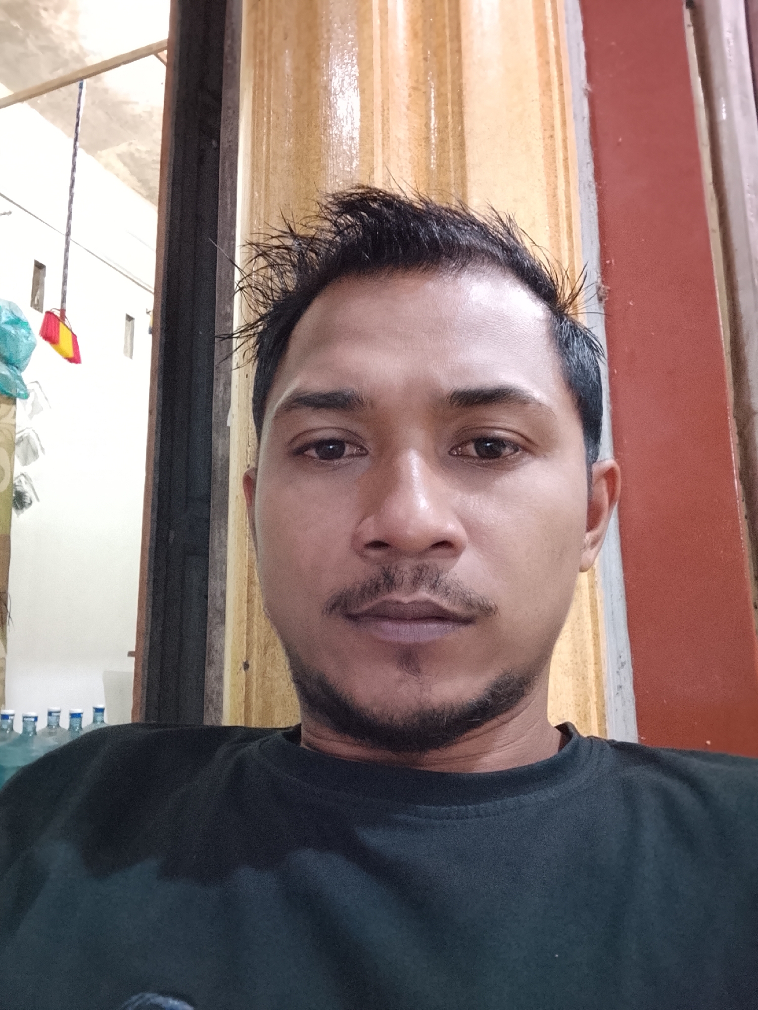 A mid-shot selfie shows a man with dark hair styled upwards and a goatee. He is wearing a black t-shirt and is positioned in front of a background that includes a vertical wooden beam, a wall with vertical yellow and red panels, and a partial view of an indoor space with some bottles and a broom in the background. The lighting suggests an indoor setting, possibly a home or a commercial establishment in Dayahlhoksukon Barat, Indonesia.