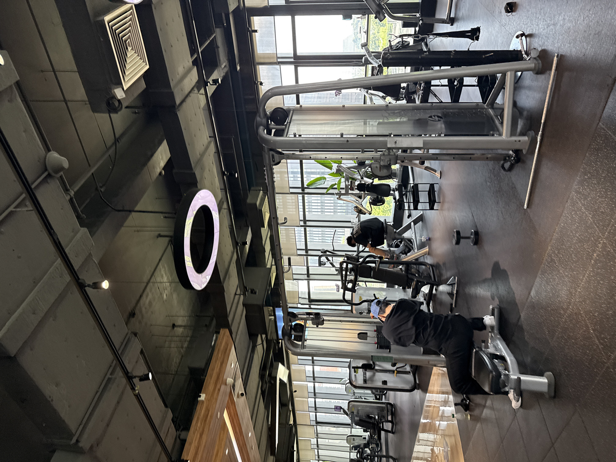 This image depicts the interior of a modern gym, likely located in Haizhu District, China, with large windows offering a view of city buildings. The scene shows rows of exercise equipment, including weight machines and benches, arranged on a dark rubberized floor. In the foreground, a person wearing a black long-sleeved shirt, black pants, and a blue baseball cap is seated on a weight bench, facing away from the camera. To the right, another individual in a black t-shirt is using a weight machine, also facing away. The gym's ceiling features exposed ductwork, track lighting, and a large circular illuminated fixture. The environment is well-lit, with natural light streaming in from the windows.