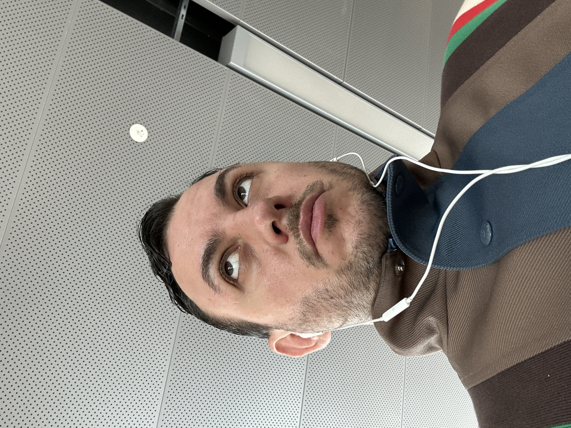 The image displays an adult male captured from a low angle, cropped from the upper chest upwards. He possesses dark hair, styled to the side, and maintains a short beard and mustache. His brown eyes are directed upwards and slightly to the right within the frame. White wired earbuds are inserted into his ears, with the wire running down his front. He is dressed in a dark blue collared garment, likely a shirt, beneath an outer jacket featuring prominent horizontal stripes. These stripes present a sequence of colors including brown, dark blue, a lighter brown, a darker brown, green, red, and cream/white. The fabric appears textured, possibly a fine rib or corduroy.
The background consists of a light gray, modular perforated ceiling, characterized by uniformly spaced small circular holes, indicative of acoustic panelling. A small, circular white object, consistent with a smoke detector or sensor, is centrally positioned on the ceiling above the subject. To the upper right, a linear, recessed light fixture with a silver-colored metallic housing is visible, running diagonally. While the architectural style of the ceiling and lighting suggests a modern indoor public space such as a contemporary transportation hub, airport terminal, or office building, the precise location within the city of London, United Kingdom, cannot be definitively identified from the visual evidence presented.