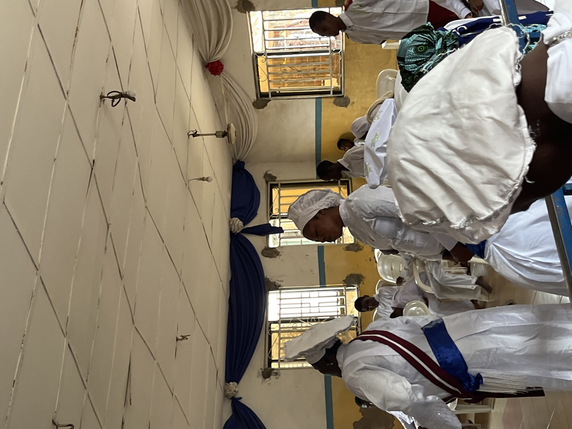 The image depicts an indoor scene, likely a religious gathering or service, taking place in a room with a suspended tiled ceiling. The ceiling has exposed wiring and a fan. Blue and white drapery adorns the upper walls. Two windows with metal bars are visible, each with yellow blinds. Several individuals, predominantly wearing white robes and head coverings, are present. Some are standing, while others are seated in white chairs. The central focus is on a woman in the foreground wearing a white robe with a blue sash and a decorative maroon and white ribbon. Her head is covered with a white cloth, and a matching white head covering is visible on another person in the foreground. In the background, other individuals, including some men and children, are also dressed in white. The overall atmosphere suggests a formal or ceremonial occasion within a modestly decorated interior space.