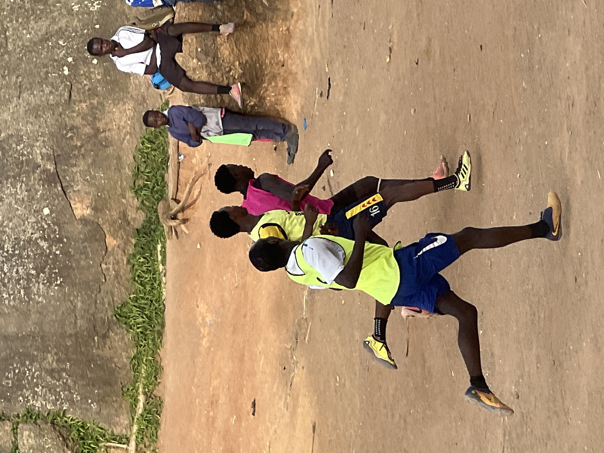 In Jos, Nigeria, three young men are engaged in a running activity on a dirt path. The man in the foreground, wearing a yellow vest over a white shirt and blue shorts with a white Nike logo, is captured mid-stride, his right leg extended behind him and his left leg pushing off the ground. He is wearing yellow and black athletic shoes. To his right, another young man in a pink vest and dark shorts is also running, his body angled forward. Behind him, a third individual in a yellow vest and dark shorts is partially visible. In the background, several other young men are seated and standing on a rocky outcrop. The environment is characterized by a dry, sandy ground with scattered debris and a large, textured rock formation in the background. Lush green vegetation is growing in patches along the base of the rock formation.