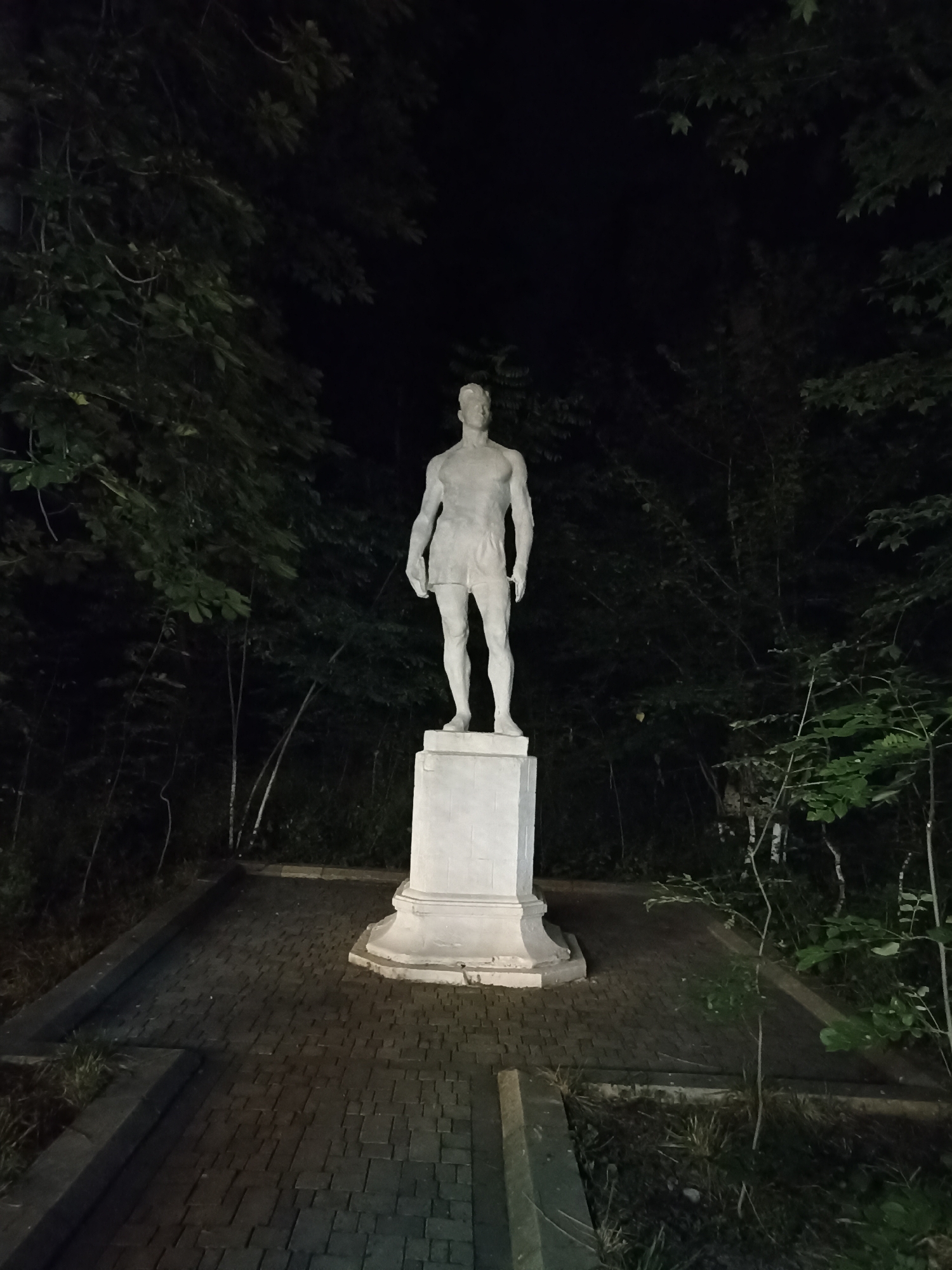 A full-shot, eye-level, nighttime photograph depicts a white, standing statue of a male figure on a white pedestal. The statue is positioned in the center of the frame, on a brick pathway surrounded by dark foliage. The statue, in a classical style, is unclothed except for shorts, with its arms at its sides and head facing forward. The pedestal is square with decorative molding. The pathway appears to be made of dark, rectangular bricks laid in a pattern. The background consists of dense trees and foliage, creating a dark, almost silhouette-like environment due to the absence of direct lighting on the natural surroundings. A single light source, likely from above and to the left, illuminates the statue and parts of the pathway. This statue is located in the city of Gusar, Azerbaijan.