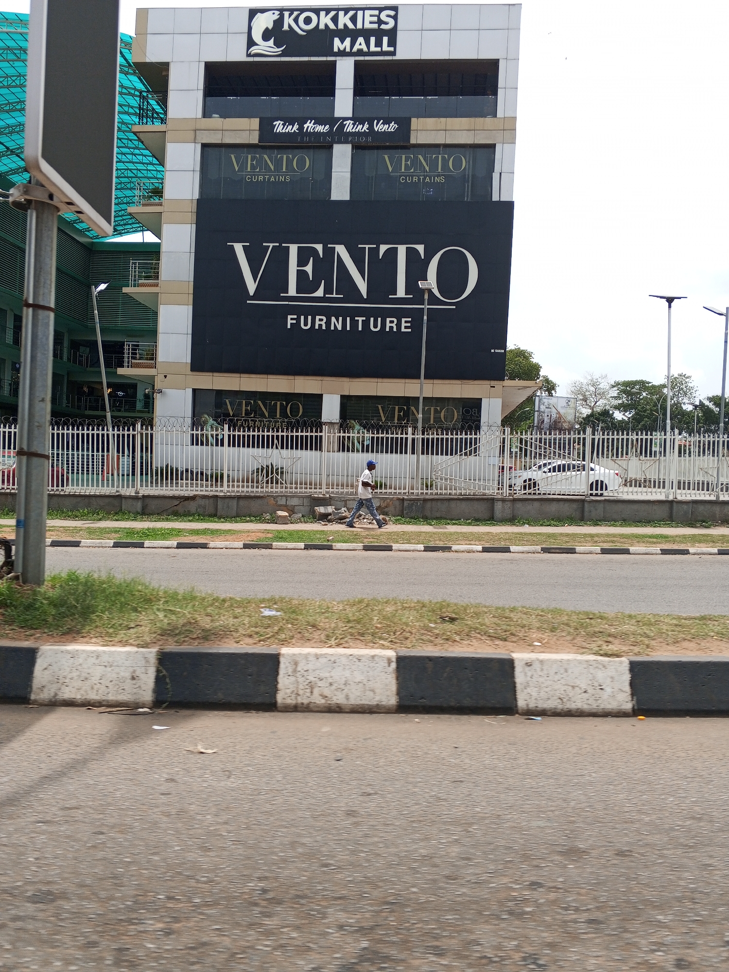 The image shows the exterior of KOKKIES Mall in Abuja, Nigeria. A multi-story building with "VENTO" branding (furniture and curtains) is visible, secured by a white metal fence along the street. A man walks on the sidewalk in front of the fence. Cars are parked inside the fence. Street lamps line the road beyond the fence. The image is taken from a low angle across a street, with a black-and-white curb in the foreground. The sky is overcast.
