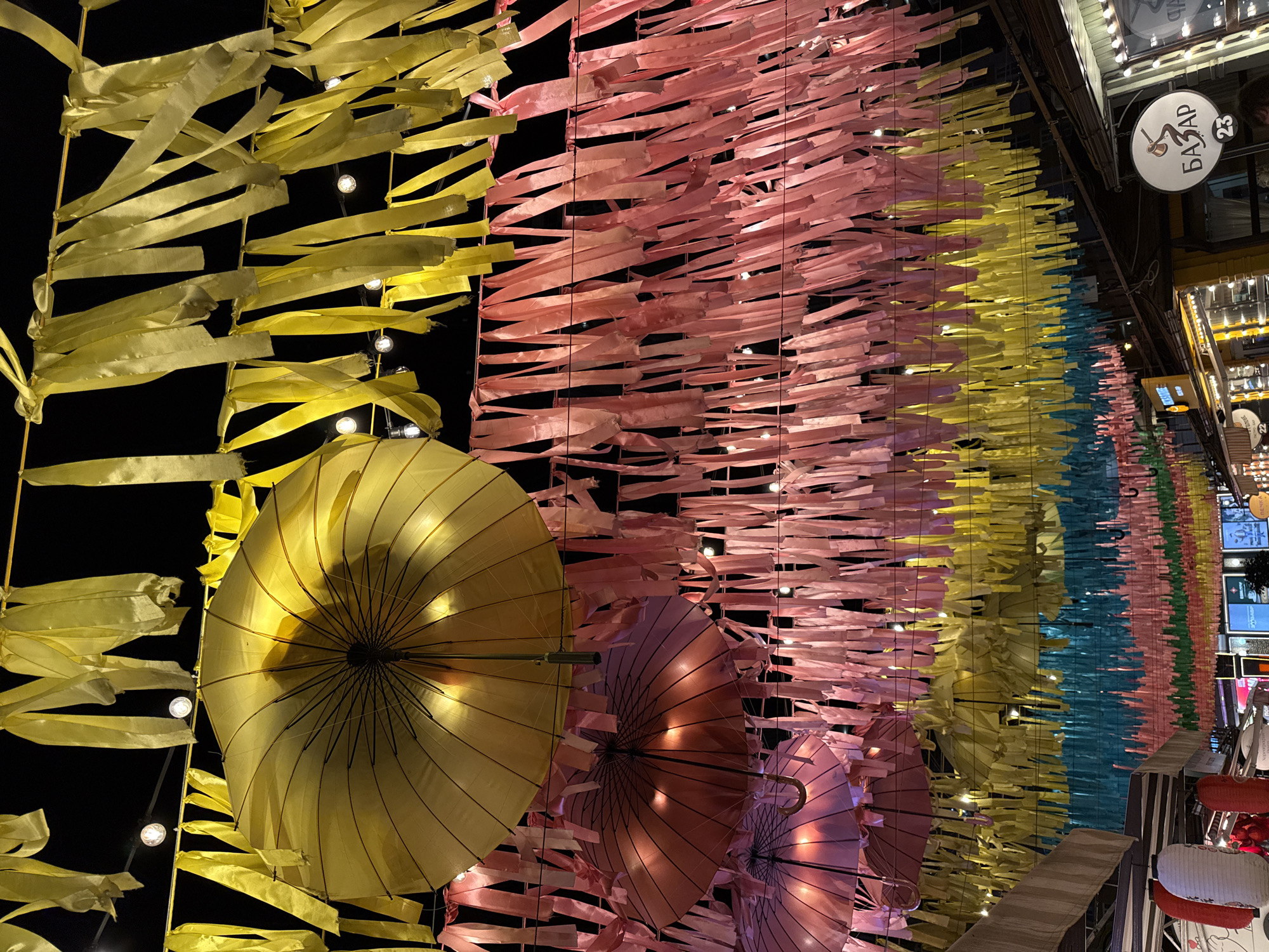 This media file captures a vibrant street scene at night, likely in Krasnodar, Russia, adorned with festive decorations. The primary subjects are a row of decorative umbrellas suspended overhead, casting a warm glow. These umbrellas, in shades of yellow and pink, are partially visible, with their spokes and fabric clearly discernible.
The scene is further enhanced by strings of colorful streamers cascading down from above. These streamers are arranged in a rainbow-like pattern, transitioning from yellow to pink, then yellow again, and finally to blue and green further down the line. Small, white lights are interspersed among the streamers, adding to the festive atmosphere.
A sign with Cyrillic text, "БАЗАР," is visible in the upper right portion of the image. This indicates a market or bazaar setting. The overall impression is one of a lively and cheerful environment, possibly a street fair or a market celebrating an event. The lighting suggests it is nighttime, and there are no indications of weather, though the decorations imply a celebratory or promotional event. The angle of the shot, looking upwards at the decorations and umbrellas, creates a sense of wonder and immersion in the festive display.