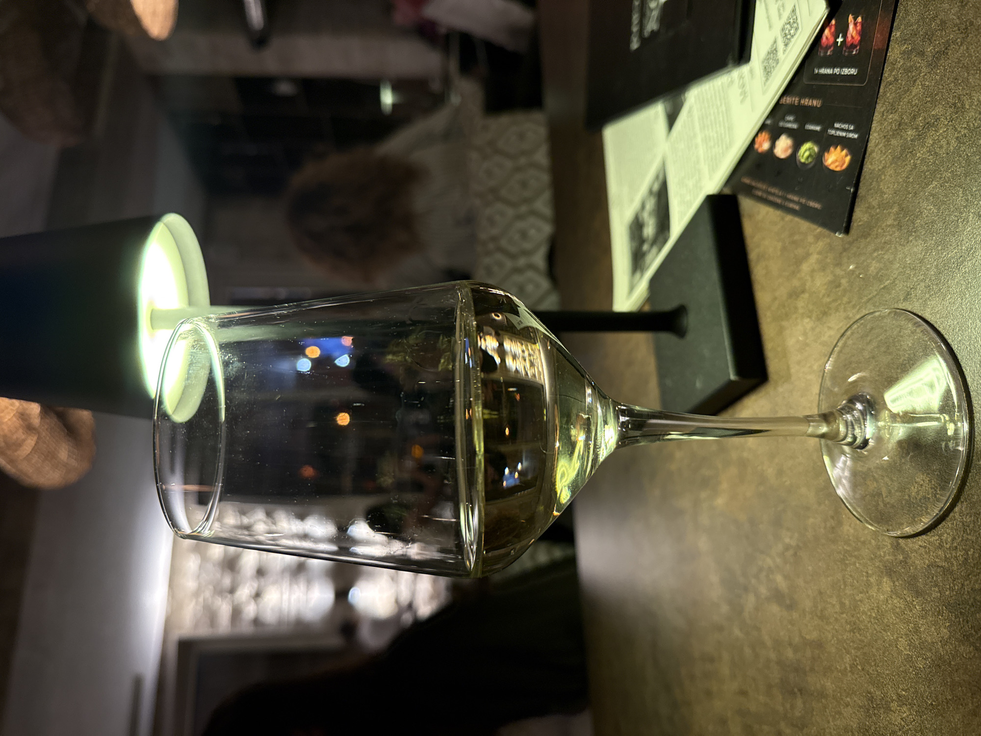 The image shows a glass of white wine on a table in what appears to be a restaurant. A menu with food and drink options is visible near the glass. The setting is in Đenovići, Montenegro, as suggested by the menu language. The environment includes a blurred background with other patrons and decorative elements, indicating a moderately busy interior space. A table lamp next to the glass illuminates the scene. No specific individuals or actions are directly observable beyond the presence of the wine glass and the setting implying a dining or drinking establishment.