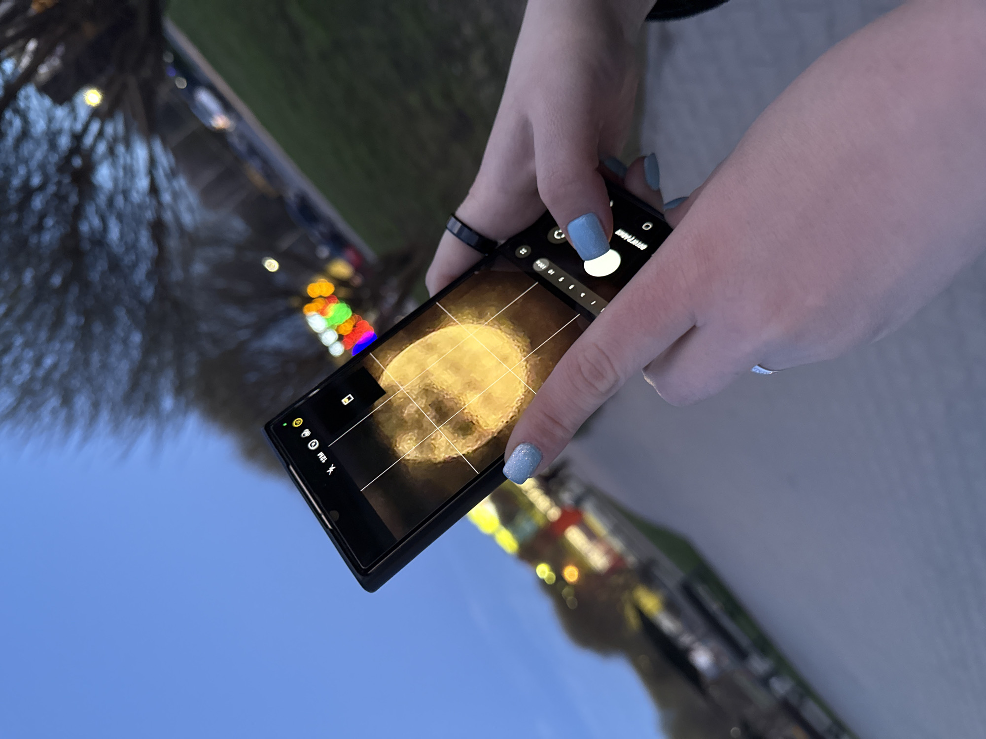 A close-up, angled shot shows a person's hands holding a black smartphone, seemingly capturing or viewing an image of the moon. The phone screen prominently displays a bright, textured full moon within its camera interface, complete with grid lines for composition.
The hands are light-skinned with light blue, glittery nail polish. One finger on the left hand wears a plain black ring, while a finger on the right hand has a silver ring with a small, clear gem. The hands are positioned as if adjusting settings or about to take a photo, with a white circular shutter button labeled "PHOTO" visible on the right side of the screen, and "MODE" above it. Other camera icons are visible on the screen's left and top edges, including a battery icon, a 'sync' or 'refresh' icon, "12M" (likely indicating 12 megapixels), and a 'starburst' icon.
The background is out of focus, suggesting a shallow depth of field. It features a deep blue sky in the lower left, transitioning to blurry dark trees or foliage. Numerous bokeh lights in warm orange, yellow, red, green, and white colors are visible in the mid-ground, implying an urban or festive environment at twilight or evening. The overall scene suggests an outdoor setting where the person is focused on photographing the moon.