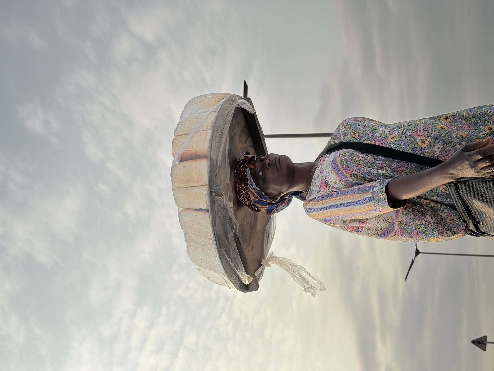 A low-angle medium shot captures a woman with dark skin wearing a colorful, floral-patterned garment and a headscarf, balancing a large circular tray of bread loaves wrapped in plastic on her head. The bread is arranged in multiple rows. The woman's face is turned slightly to her right, her gaze directed forward and upward. A dark strap bag is slung across her right shoulder. The background is a pale, overcast sky filled with wispy clouds. Two streetlights are visible in the distance, one behind and to the right of the woman, and another further to the left. A triangular traffic sign is also present in the lower left corner of the frame. The scene appears to be outdoors, possibly on a street in Alaguntan, Nigeria.
