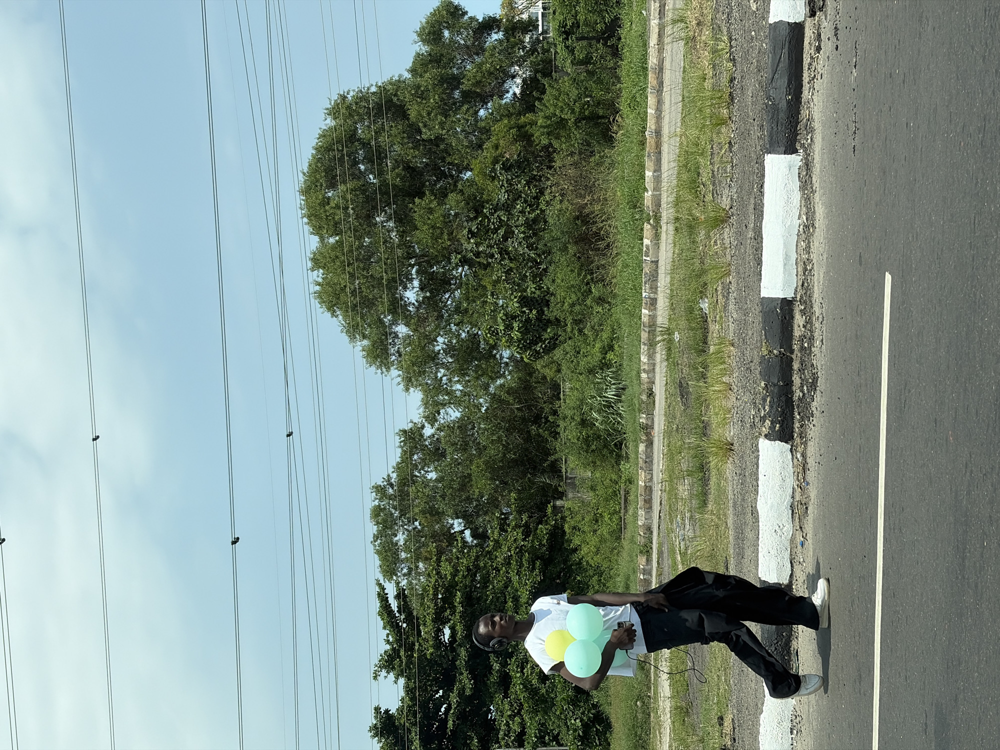 A person stands on the side of a road in Ojomu, Nigeria. The individual, wearing a white t-shirt, black pants, and headphones, holds several balloons. A black jacket hangs over their shoulder. To the side of the road is grassy vegetation. Overhead power lines cross the sky. A curb is painted with black and white stripes.