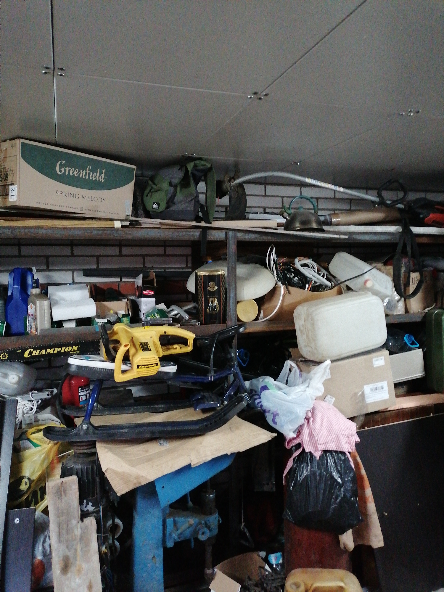 The image depicts a cluttered storage area, possibly a garage or basement, filled with various objects and materials. Shelves line the walls, laden with boxes, containers, and miscellaneous items. A "Greenfield Spring Melody" box is visible on the upper shelf. Below, a yellow "Champion" chainsaw sits among other tools and equipment. A blue piece of machinery is partially visible towards the bottom of the frame. A black garbage bag and other containers are visible on the lower level. The specific location is unknown as no city or location is specified.