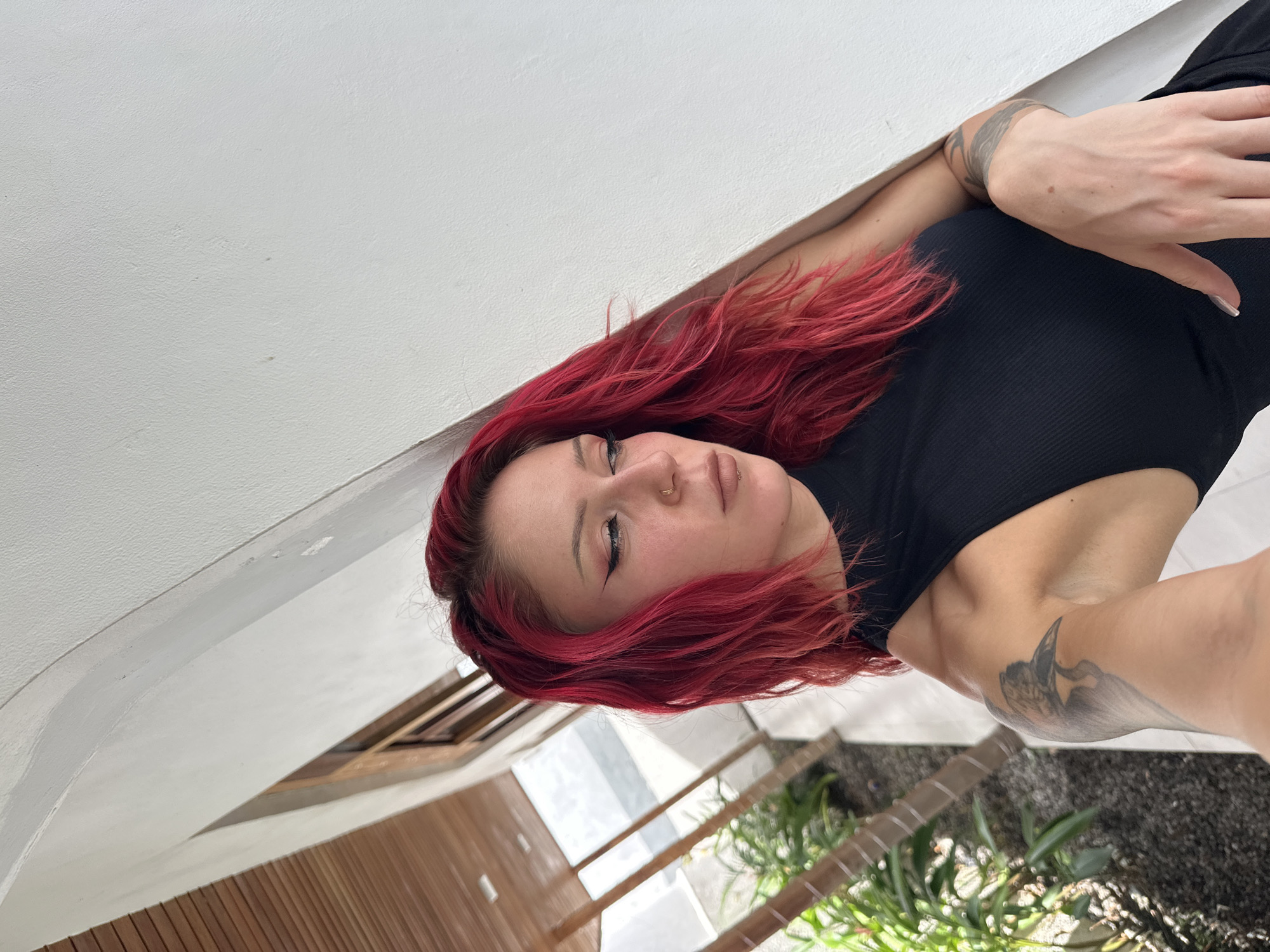 A medium shot captures a woman with dyed red hair and fair skin lying on a white surface, likely a wall or ceiling. She is wearing a black top. Tattoo art is visible on her arms. The background suggests an outdoor or semi-outdoor setting with wooden structures, possibly a building facade. Green plants are also in the frame, pointing to a natural or garden-like setting. Based on the location details in the prompt, the scene is likely in a residential or commercial area of North Kuta, Indonesia, possibly in a building with open-air or garden features.