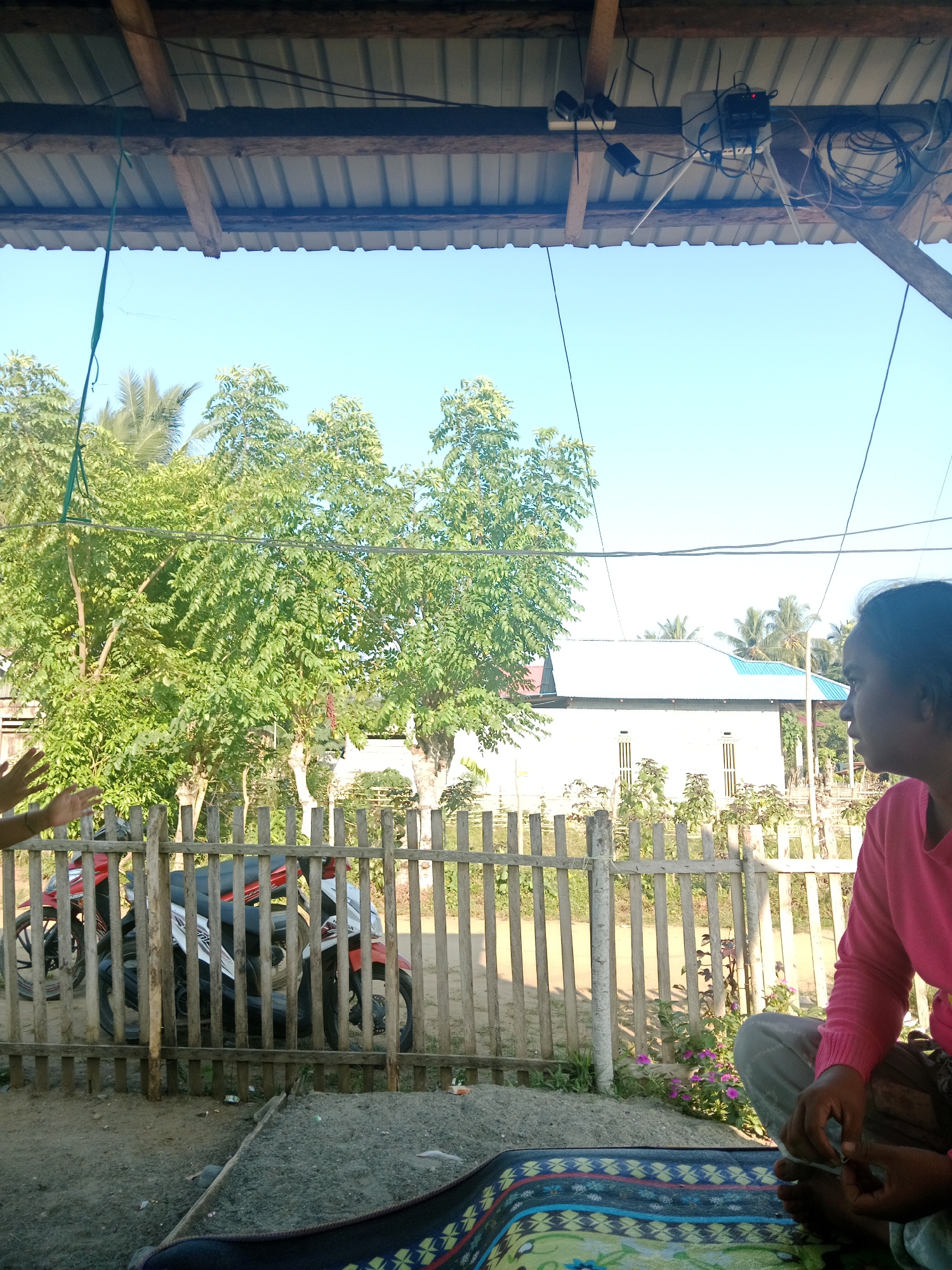 In Lakea Satu, Indonesia, a scene unfolds outdoors under a covered structure with a corrugated metal roof supported by wooden beams. The sky is clear and blue. In the foreground, to the right, a person wearing a pink sweater and grey pants is seated on a colorful mat. Their hands are visible, with one holding a cigarette. To the left, another person's hands are raised. Beyond them, a wooden fence encloses a small area where two motorcycles, one red and white, are parked. Behind the fence, lush green trees and foliage are abundant, with palm trees visible in the distance. A white building with a blue-tiled roof stands behind the trees, featuring several windows with light-colored shutters. Electrical wires and what appear to be network equipment are suspended from the roof structure above. The ground in the foreground consists of sand and dirt, with some debris scattered around.