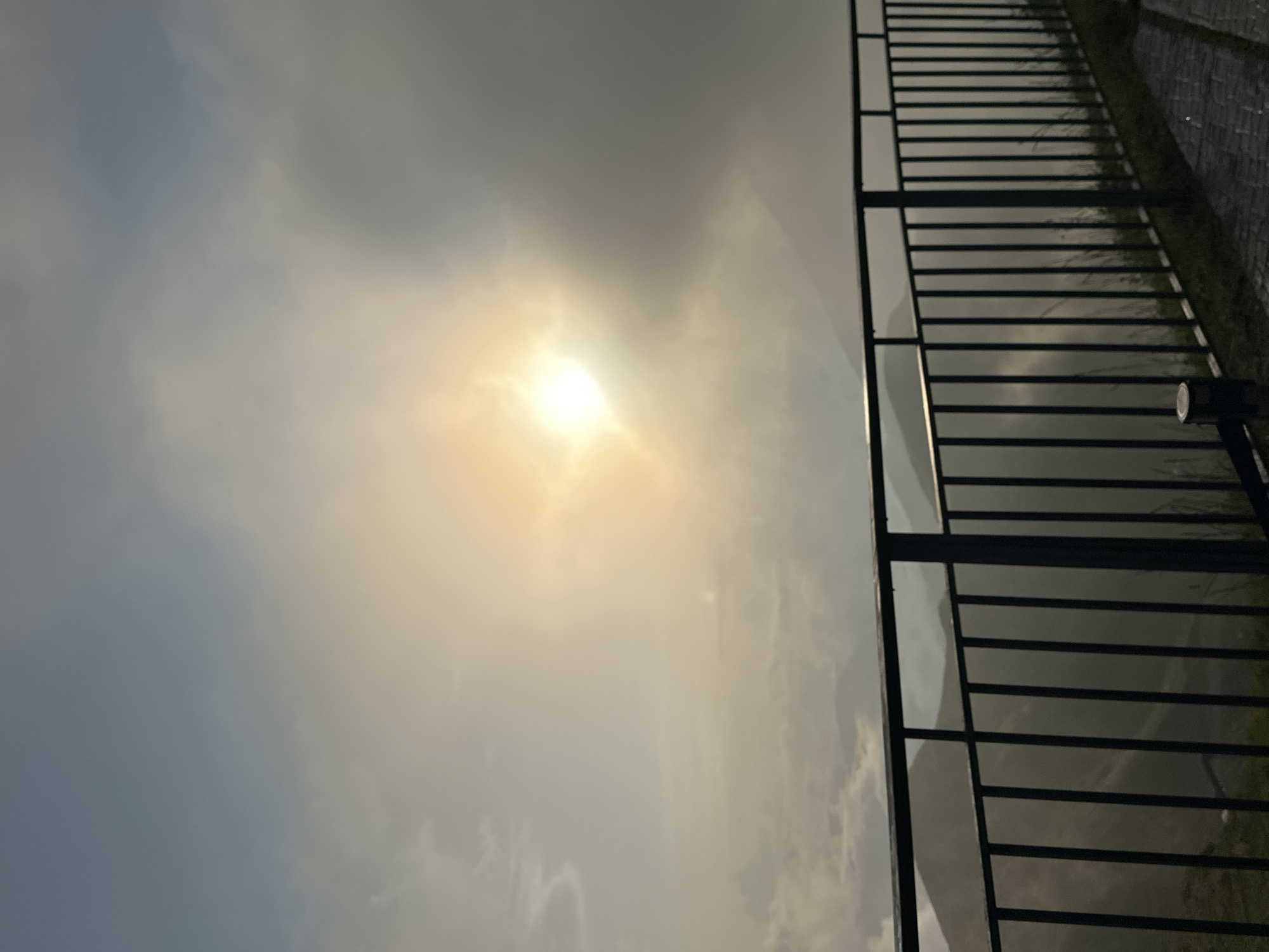 The image depicts a scenic view of mountains shrouded in mist, viewed from behind a dark metal railing. The sun is partially visible through the thick clouds, casting a warm, diffused glow on the scene. The sky transitions from a pale blue at the top to a hazy yellow and gray near the horizon. In the foreground, the railing runs horizontally across the lower portion of the frame, with vertical bars spaced evenly. Beyond the railing, layers of mountains fade into the distance, their shapes softened by the atmospheric conditions. A paved walkway with brick-like patterns is visible to the right of the railing, suggesting this is a designated viewpoint or observation deck. The environment is natural and mountainous, characteristic of a location like Estosadok, Russia, known for its alpine scenery and ski resorts. There are no people or animals visible in the frame.
