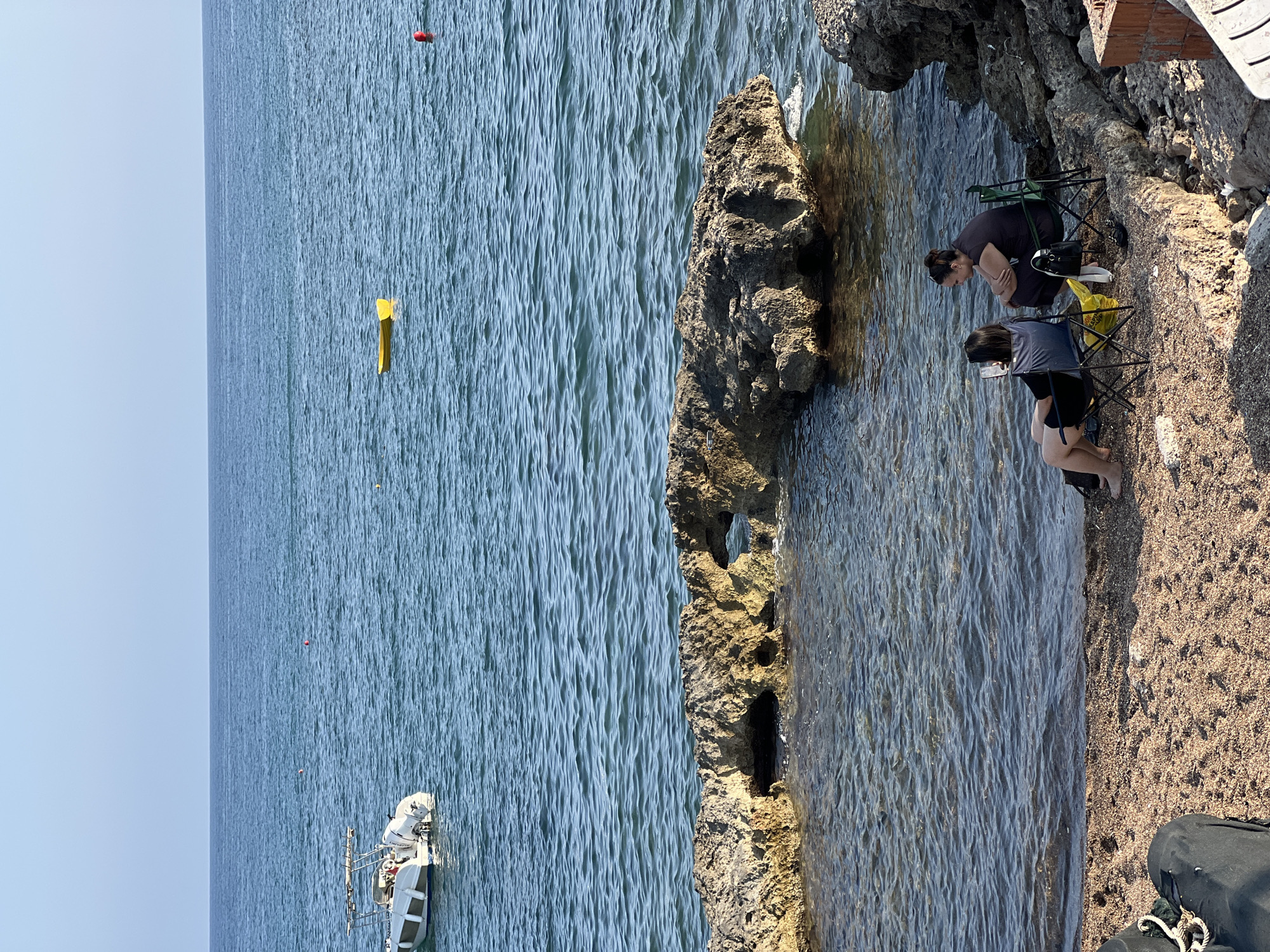 Two individuals are seated on beach chairs on a pebble-and-sand shore in Antalya, Turkey, with a rocky outcrop to their right and the clear blue Mediterranean Sea before them. The individual on the left is looking at a mobile phone, while the person on the right is looking towards the sea. A yellow flotation device and a boat are visible in the water. The sky is clear and blue.