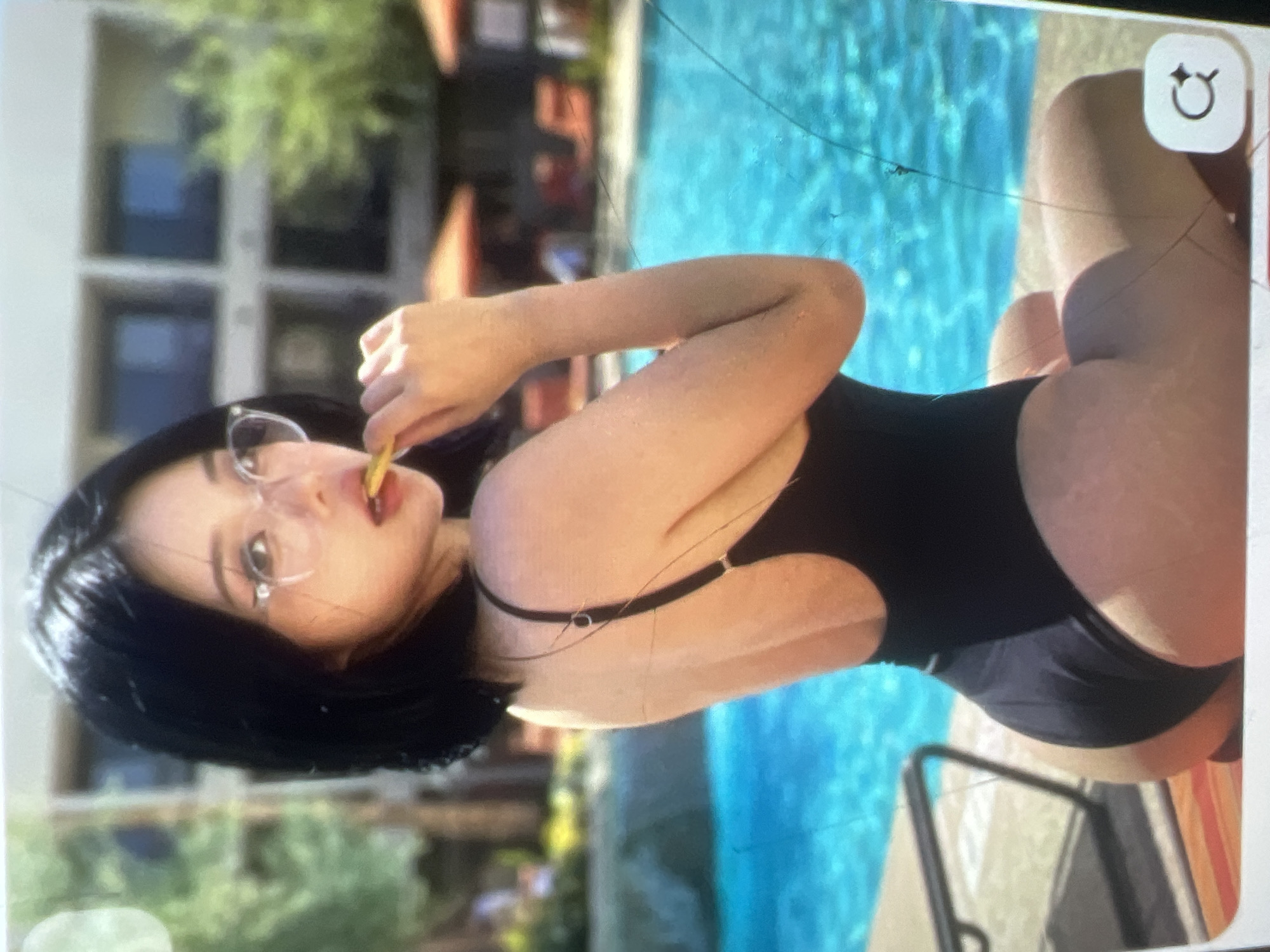 The image shows a young woman wearing a black one-piece swimsuit and glasses. She is sitting by a swimming pool, with her back to the camera. She is holding a yellow object, possibly a piece of candy, in her mouth. The background shows a building and trees. There is no information available to determine the location of this image in the city of Kara-Dıykan, Kyrgyzstan.