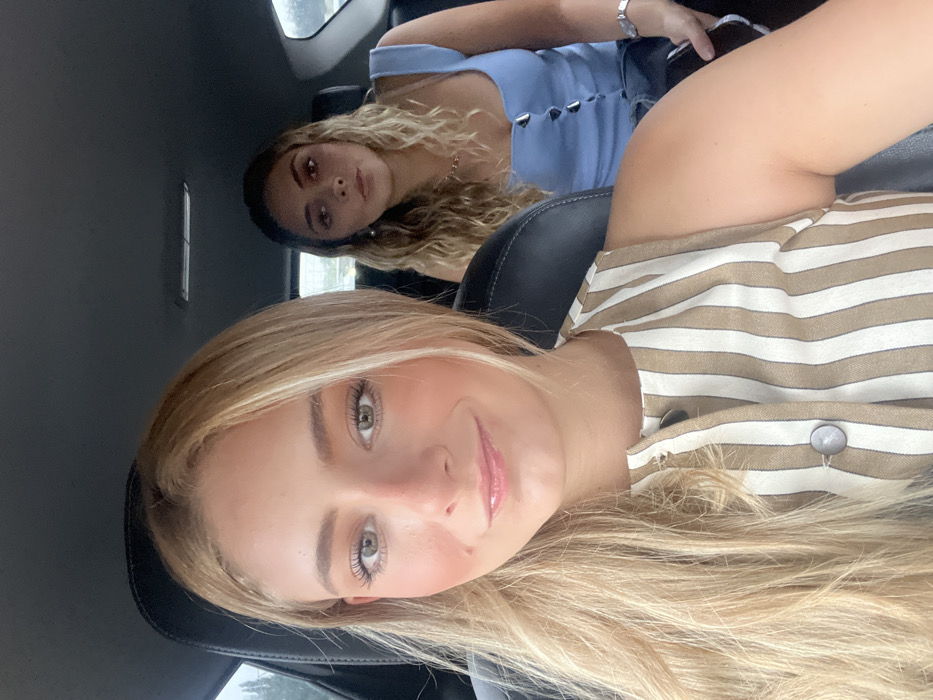 This image depicts two young adult females inside the passenger cabin of a vehicle, located in Camargo, Spain. The female in the immediate foreground, positioned in what appears to be the front passenger seat, faces the camera directly with a slight smile. She has long, light blonde hair parted centrally and light-colored eyes, wearing a sleeveless garment with vertical stripes of beige and off-white featuring a buttoned placket. The second female is situated in the rear passenger seat behind the first, her head slightly turned to her right, not directly engaging the camera. She has wavy, light-toned hair and is wearing a light blue sleeveless top with a buttoned front and a silver-toned wristwatch on her left wrist. The vehicle's interior consists of dark-colored seating surfaces. Through the windows, a blurred outdoor environment with green foliage is visible.