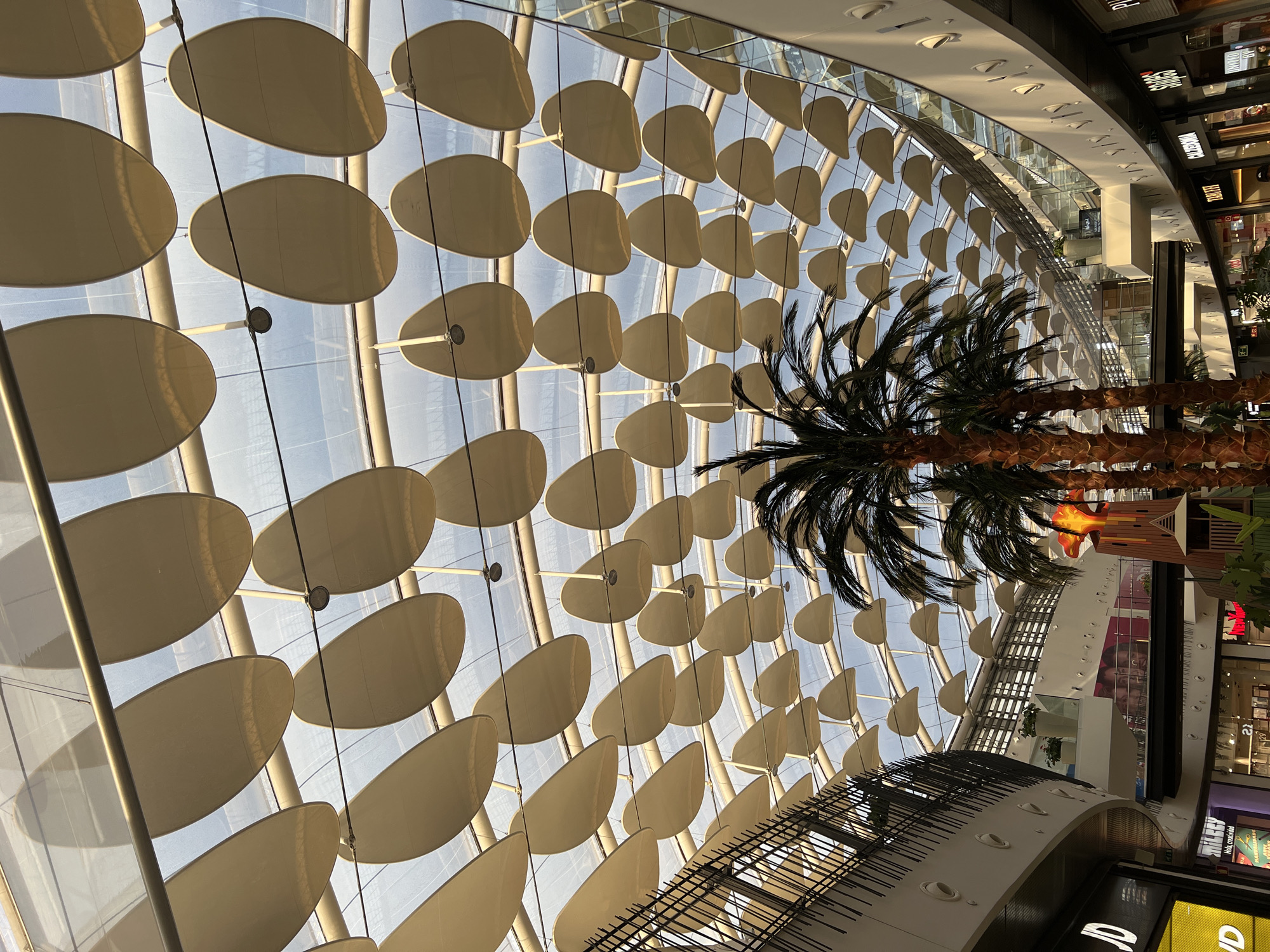 The image shows the interior of a shopping mall with a distinctive domed glass ceiling featuring numerous oval-shaped fabric panels suspended below. The panels are arranged in a grid-like pattern and cast soft shadows. Natural light streams through the glass roof, illuminating the scene. In the foreground, several artificial palm trees are visible, adding a tropical feel to the environment. The mall appears to be multi-level, with balconies and shops visible on different floors. Some shop signs are partially visible, including "JD" and "CHILLY". A decorative structure resembling a volcano or tower is also present. The architecture suggests a modern design. The specific location is identified as the "Plaza Río 2" shopping center in Madrid, Spain. There are no visible people in the frame. The overall impression is of a bright, spacious, and architecturally interesting indoor public space.