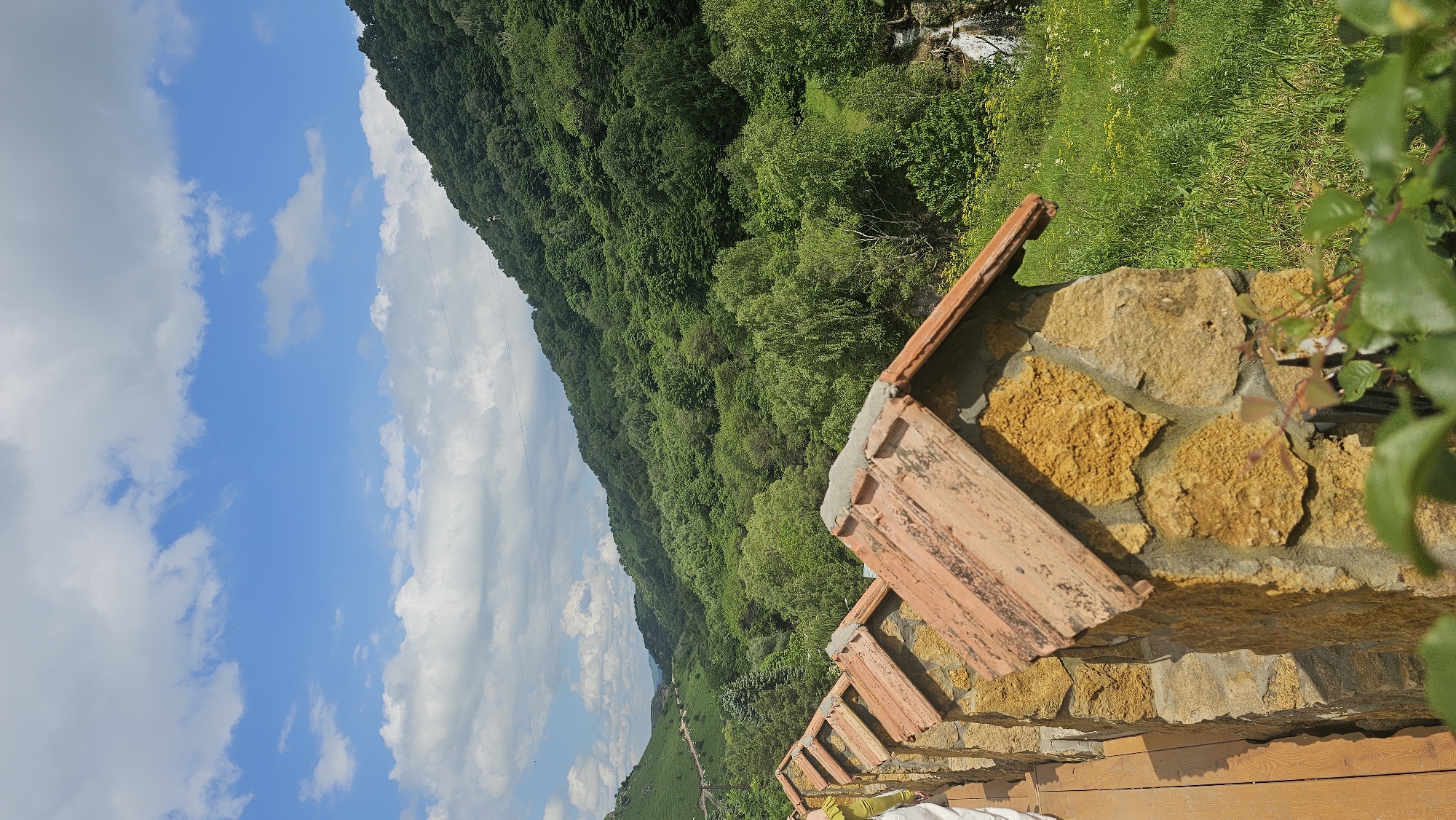 The image depicts an outdoor scene with a panoramic view of a lush green mountainside under a bright blue sky with scattered white clouds. In the foreground, there is a stone wall with terracotta roof tiles extending along its top. The wall is constructed from irregularly shaped, sandy-colored stones, mortared together. Some of the roof tiles show signs of wear and age. To the right of the wall, a wooden structure is visible, possibly a deck or railing. Green foliage, including some branches with leaves, frames the right side of the image.
The mountainside is densely covered with trees, showcasing a variety of green hues, suggesting a vibrant summer landscape. A faint path or road can be seen winding its way up the lower part of the mountain. In the upper right portion of the image, a small waterfall or stream is visible cascading down the slope amidst the greenery.
The overall impression is one of natural beauty and a tranquil outdoor setting. The lighting suggests it is daytime, likely late morning or early afternoon, with clear weather. No people or specific text are clearly discernible in the image. The landscape evokes a sense of peace and immersion in nature. The location context provided is Krasny Kurgan, Russia.