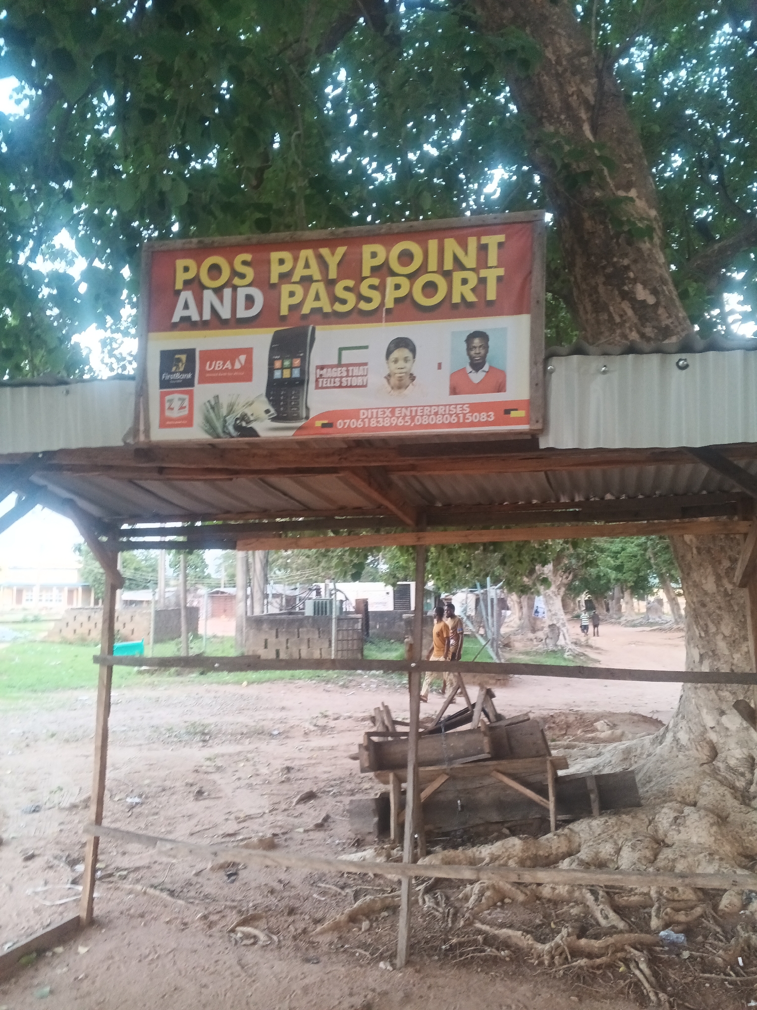 This media file is a photograph of an outdoor stall with a sign indicating services offered. The sign reads "POS PAY POINT AND PASSPORT" and displays logos for FirstBank and UBA, suggesting these are supported payment methods. Below this, it mentions "DITEX ENTERPRISES" with phone numbers. Images on the sign include a POS machine, a stack of banknotes, and headshots of two individuals, one female and one male. The stall itself is constructed with corrugated metal roofing and wooden supports.
The scene appears to be in a somewhat dusty, rural or semi-urban environment, possibly in Jalingo, Nigeria, as indicated by the location context. The ground is dirt, with some scattered debris and tree roots visible. In the background, there are other structures, some made of brick, and a few people are visible walking in the distance. The lighting suggests it is daytime, and the presence of lush green trees indicates a possibly warm climate. The overall atmosphere is casual and functional, representing a local service point. There are no discernible emotions expressed by the people in the image, who are at a distance.
