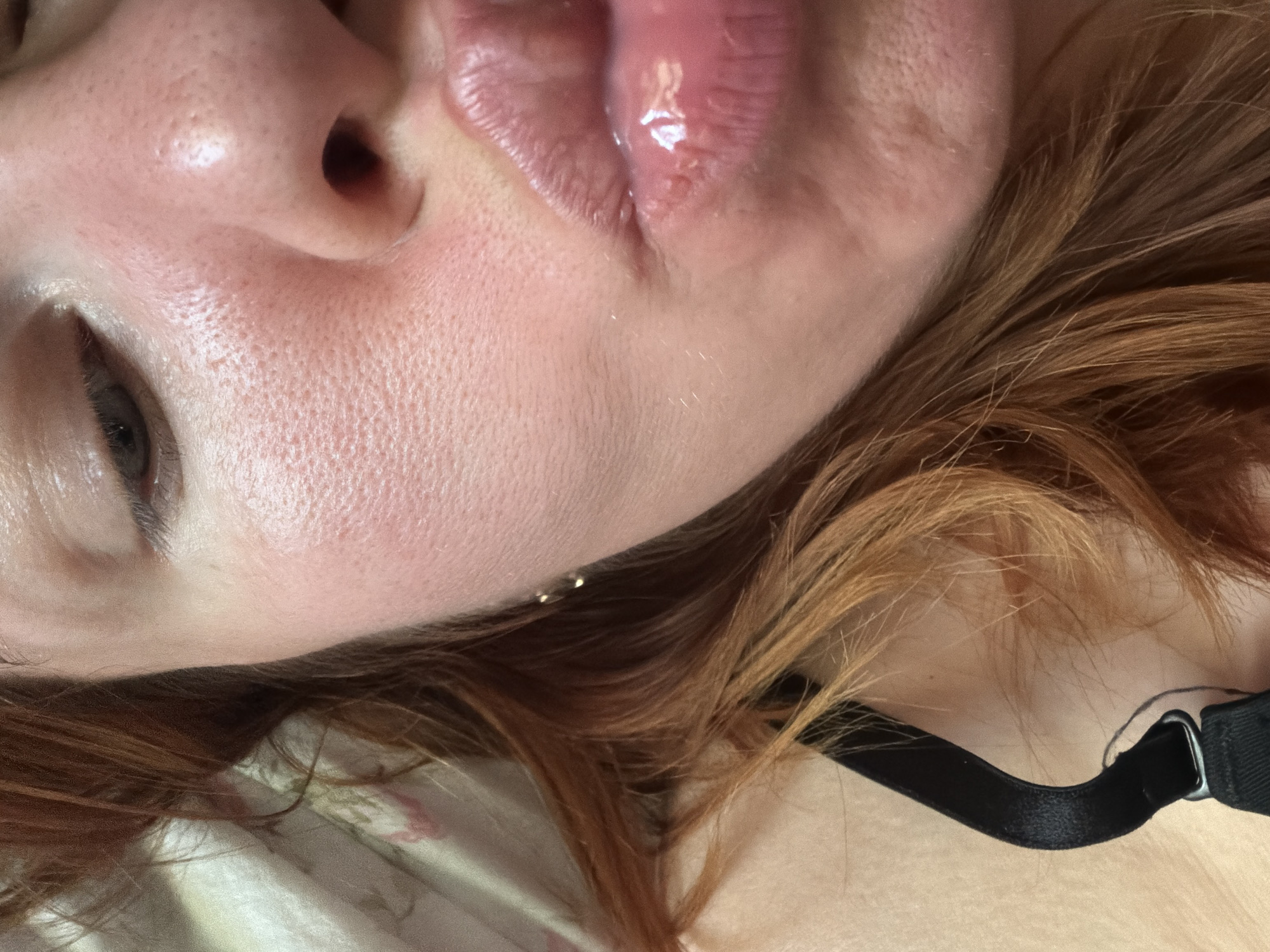The image is a close-up, low-angle shot of a person's face, focusing on the cheek, nose, and lips. The person has fair skin with visible pores and a reddish tone. Their lips are glossy and appear to be slightly pursed. Reddish-brown hair is visible on the left side of the frame, and a black bra strap is also present in the lower left corner. The background is blurred and appears to be a patterned fabric. The image does not contain any elements that can be used to determine a location in the city of Podolsk, Russia.