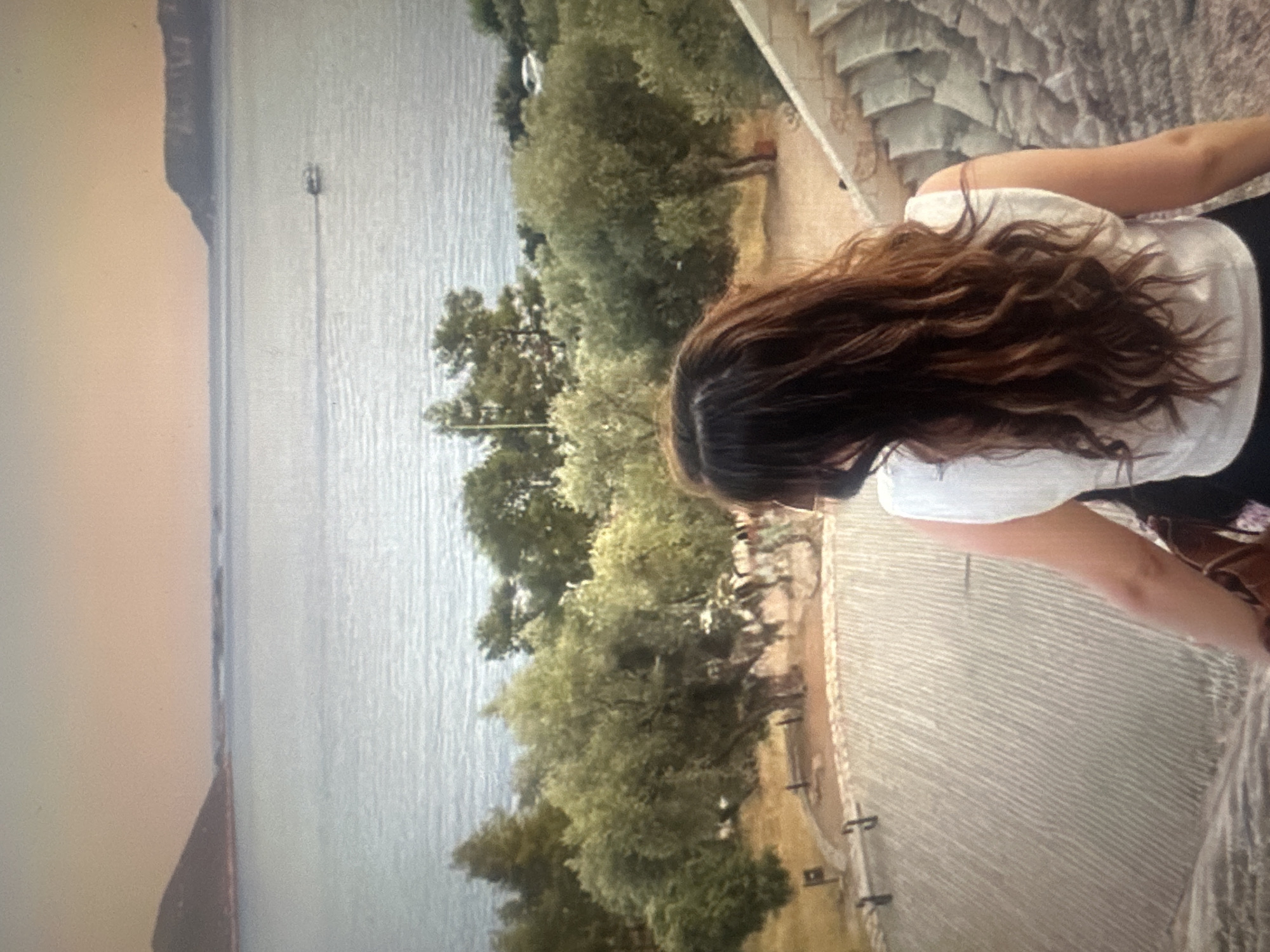 A woman with long, wavy brown hair stands on a stone structure, facing away from the camera and looking out at a vast body of water. She is wearing a white sleeveless top and a brown bag is visible over her right shoulder. The water is calm and reflects the soft, hazy light of what appears to be sunset or sunrise, with a warm, orange-tinted sky above. In the distance, land masses are visible on the horizon, with hills or mountains on either side. A small boat is making a wake on the water, heading towards the right side of the frame. In the middle ground, there are several large, mature trees with dense green foliage. The stone structure the woman is standing on has a textured, stepped appearance. The ground in front of the trees is a reddish-brown color and appears to be a pathway. The overall scene evokes a sense of tranquility and scenic beauty.