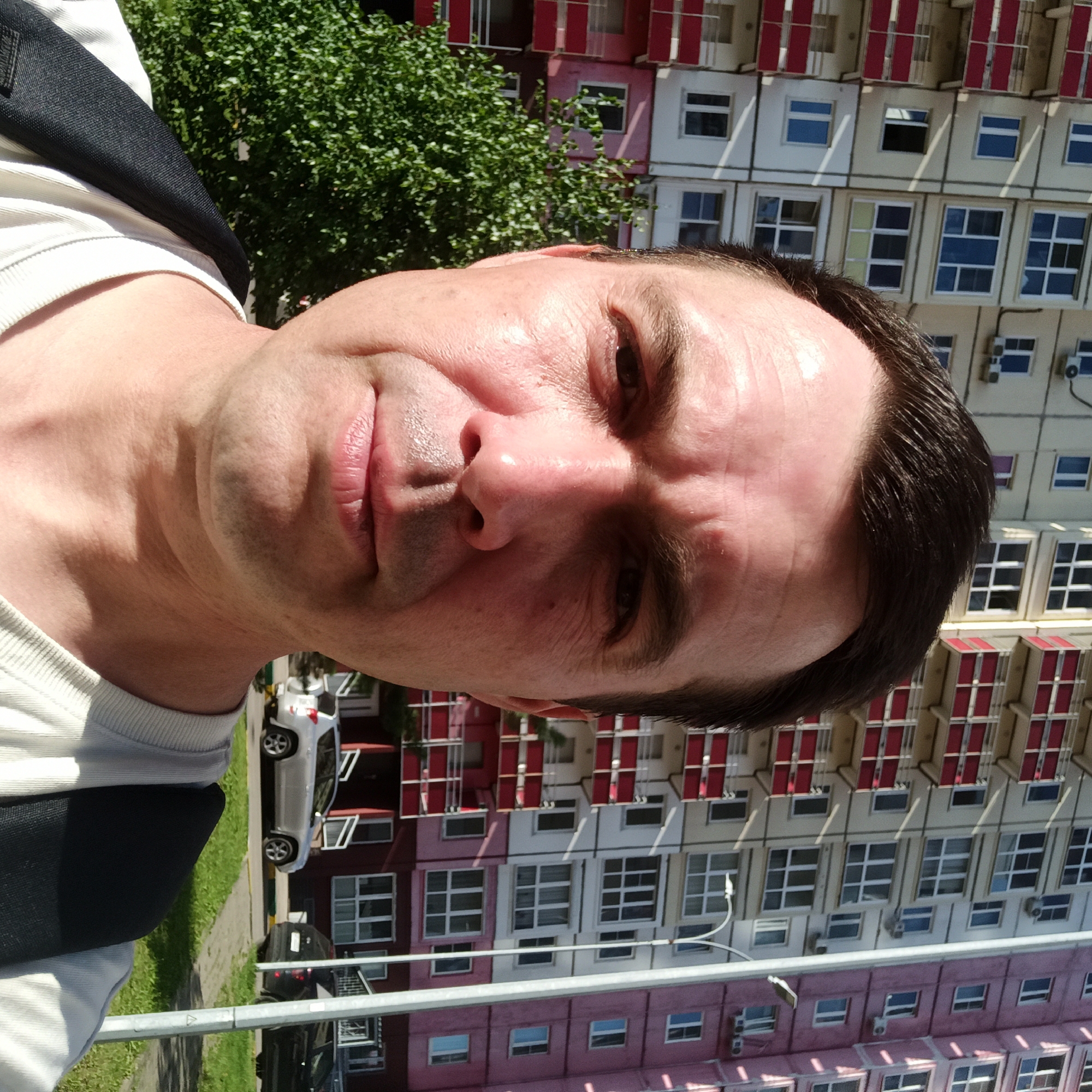 A close-up, selfie-style, medium shot captures a man in his late 30s to early 40s with dark hair, a light complexion, and fair skin. He is positioned slightly off-center to the left, facing the camera directly with a neutral expression. His head and shoulders are the primary focus, with the background showing a tall, multi-story apartment building under a bright, sunny sky. The building exhibits a pattern of balconies with red railings and beige and pink painted sections. The man is wearing a light-colored t-shirt and a dark backpack. To his left, a lush green tree provides a natural contrast to the urban architecture. Further in the background, a silver SUV is parked in front of the building, with a glimpse of a street and sidewalk. The image appears to be taken outdoors in a residential area of Nizhny Novgorod, Russia.